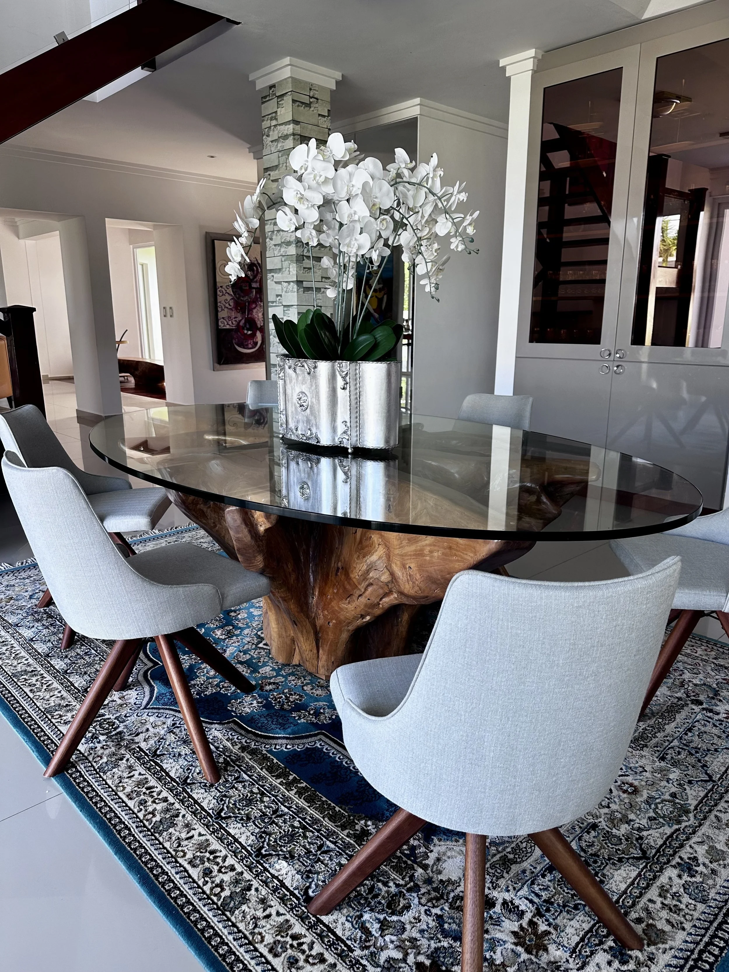 Modern dining room with a wooden and glass table, common area with chairs, a decorative rug, and a plant arrangement with white flowers in a silver vase.