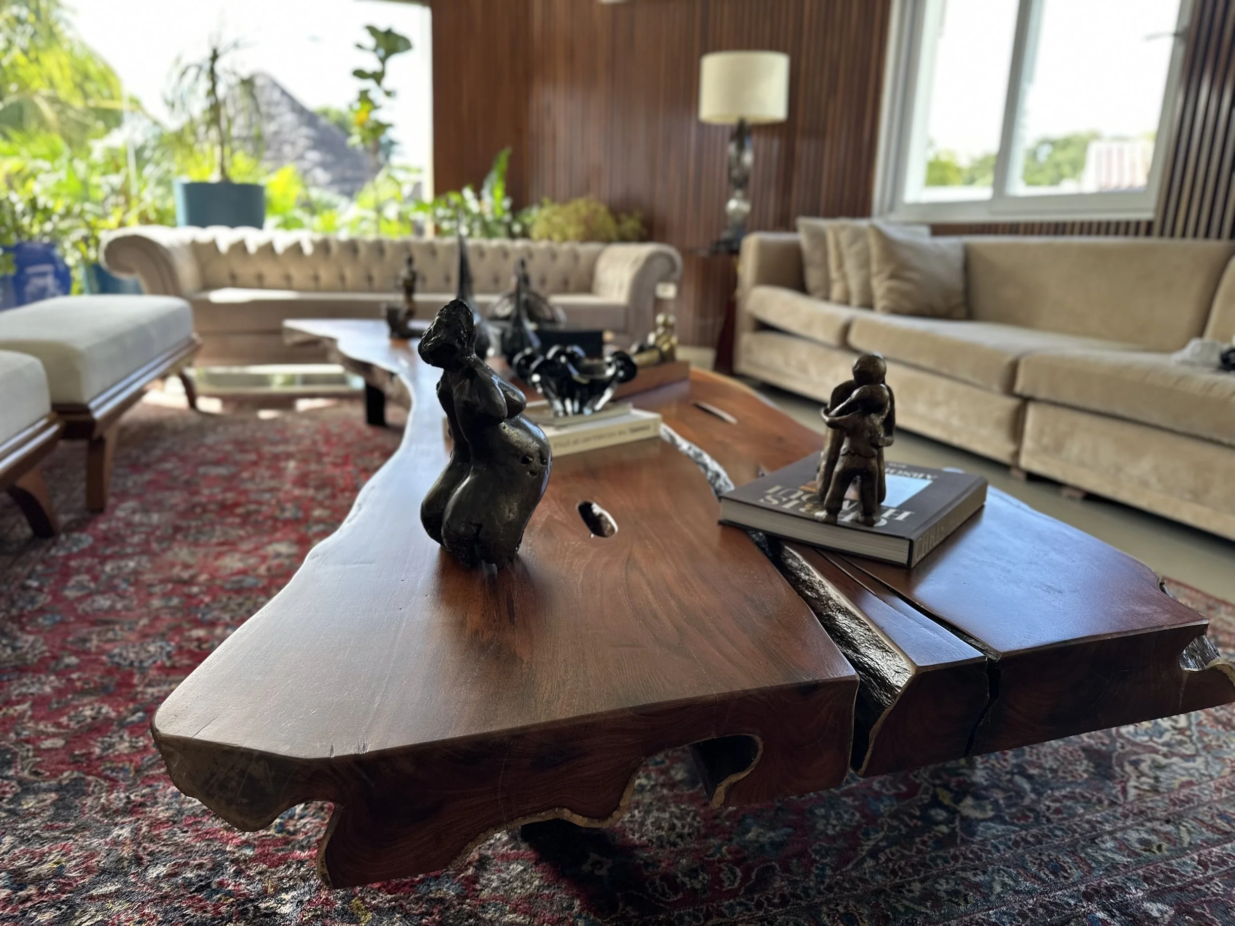 Living room with wooden coffee table, beige sofas, lamps, and large windows with greenery outside.