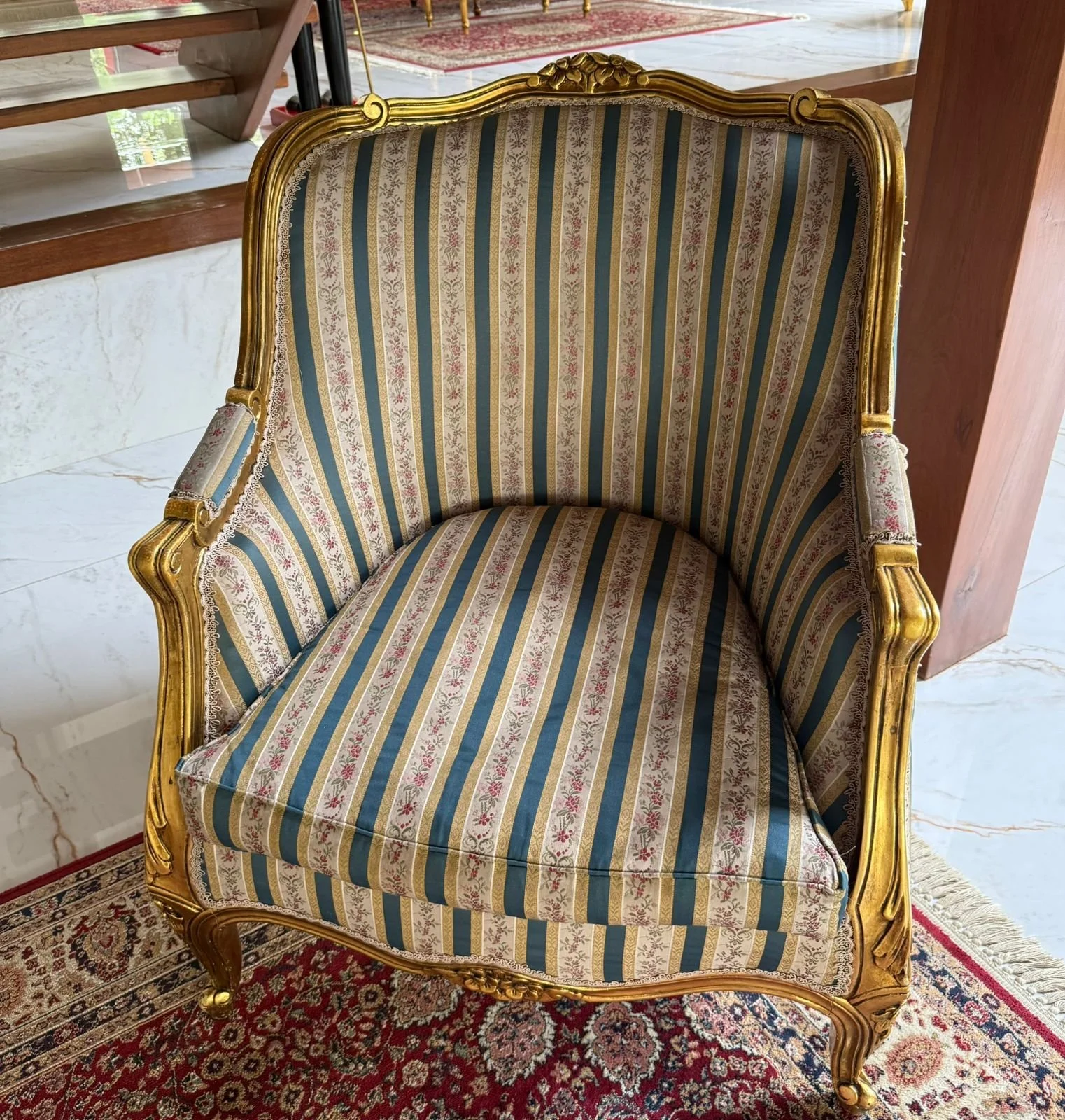 A vintage armchair with a gold ornate wooden frame and striped upholstery in beige, blue, and gold, placed on a patterned rug.
