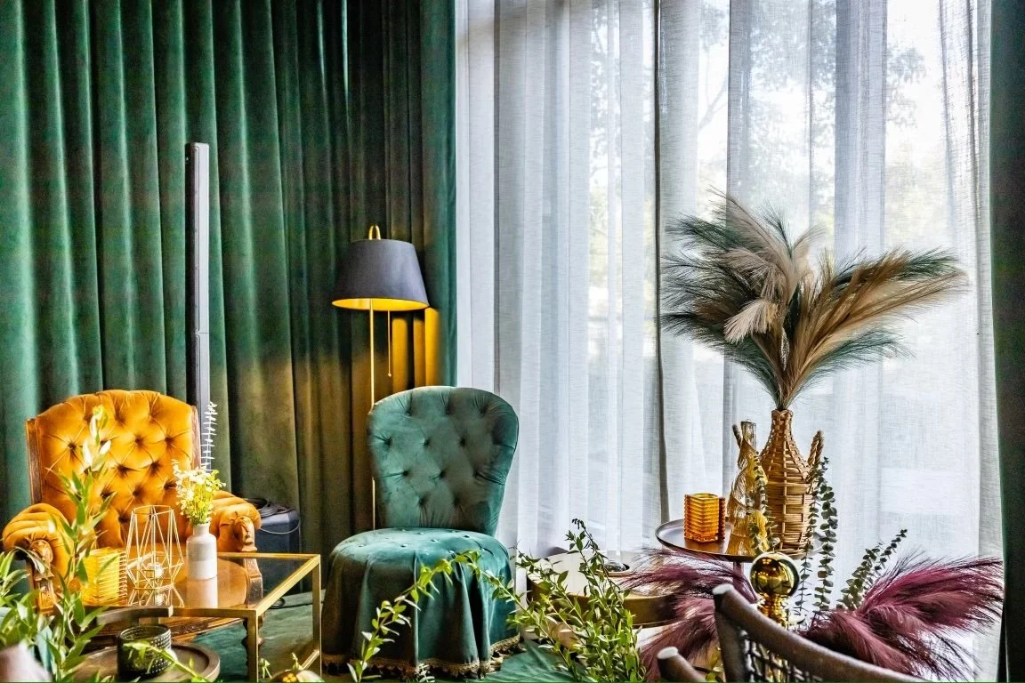 Living room with green curtains, a gold and glass side table, a yellow tufted armchair, a green tufted armchair, a black floor lamp, decorative vases with pampas grass, and various ornamental plants.