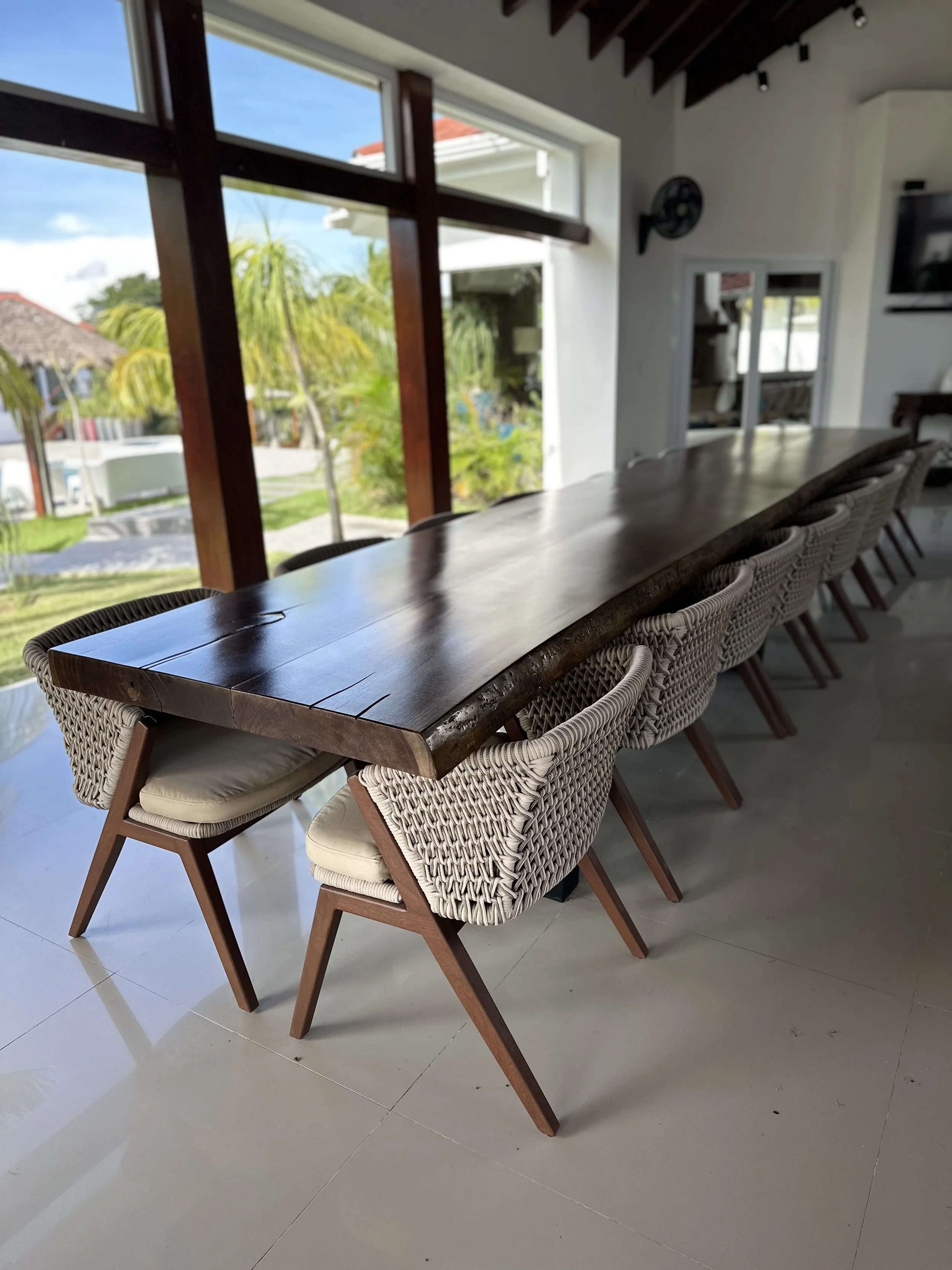 Long wooden dining table with woven rattan chairs inside a bright room with large windows showing a tropical outdoor scene.