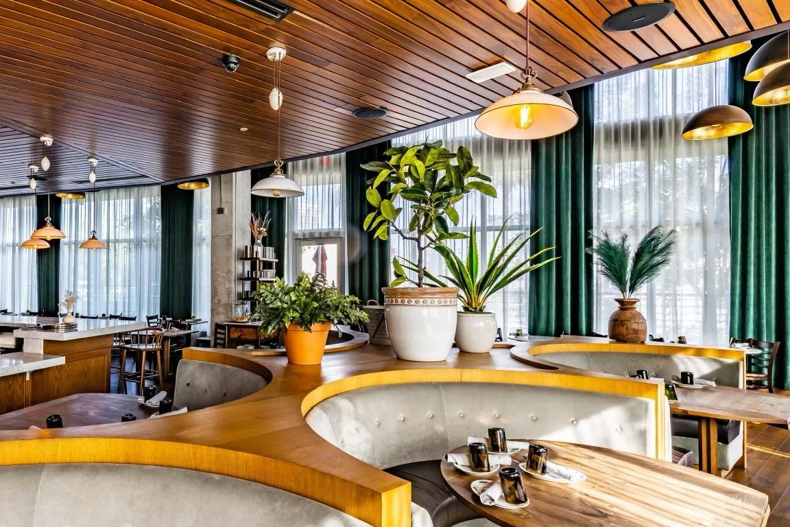 A restaurant interior with round wooden tables, upholstered seating, and potted plants near large windows with curtains, decorated with pendant lights hanging from a wood-paneled ceiling.