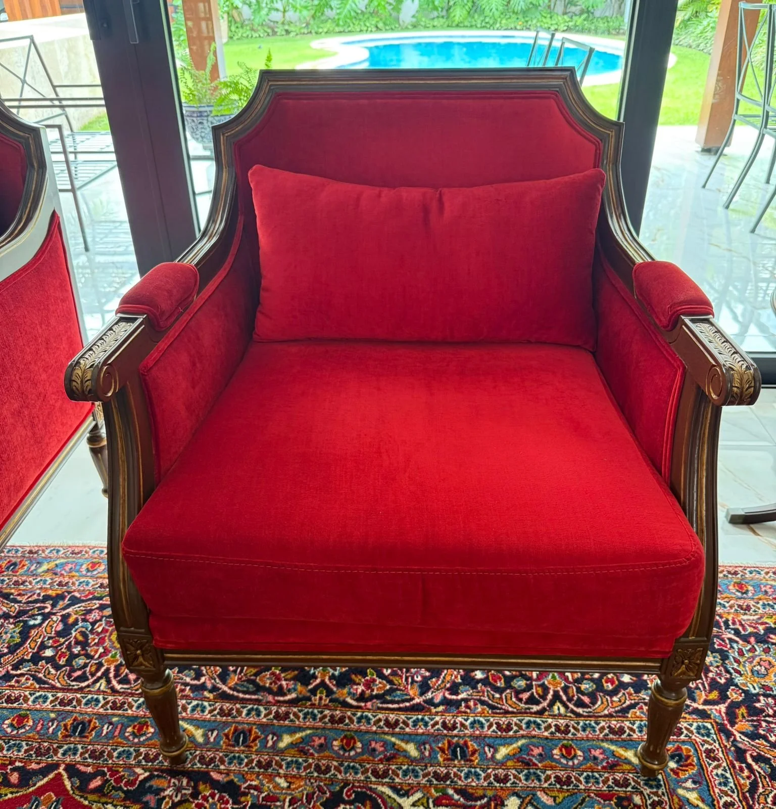 Red upholstered vintage armchair with carved wooden frame and cushion on a patterned rug, near glass doors leading outside to patio and swimming pool.