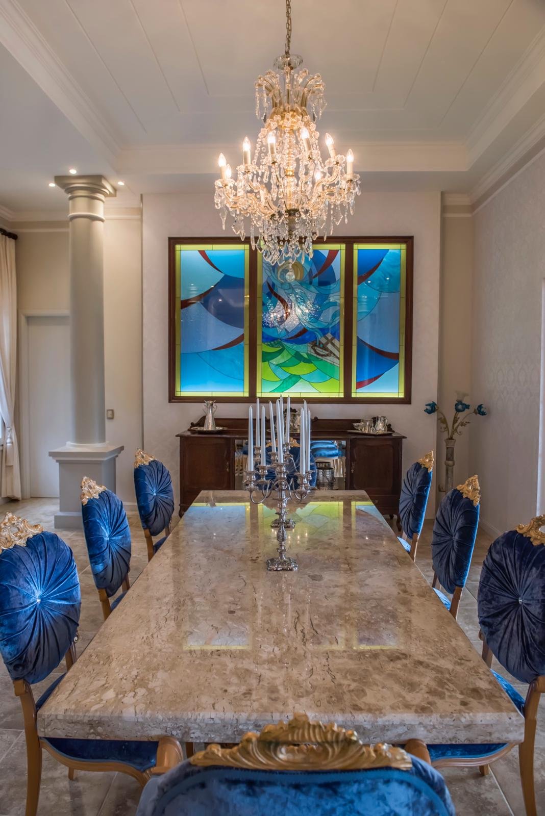 Elegant dining room with a large marble table, ornate gold and blue chairs, a chandelier hanging above, and a colorful stained glass window in the background.