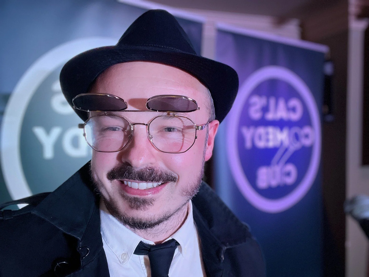 A man smiling, wearing a black hat, glasses with one lens flipped up, a white shirt, black tie, and black jacket, with a backdrop of signs reading 'CALS' and 'GAY'. the image appears to be indoors with purple lighting.