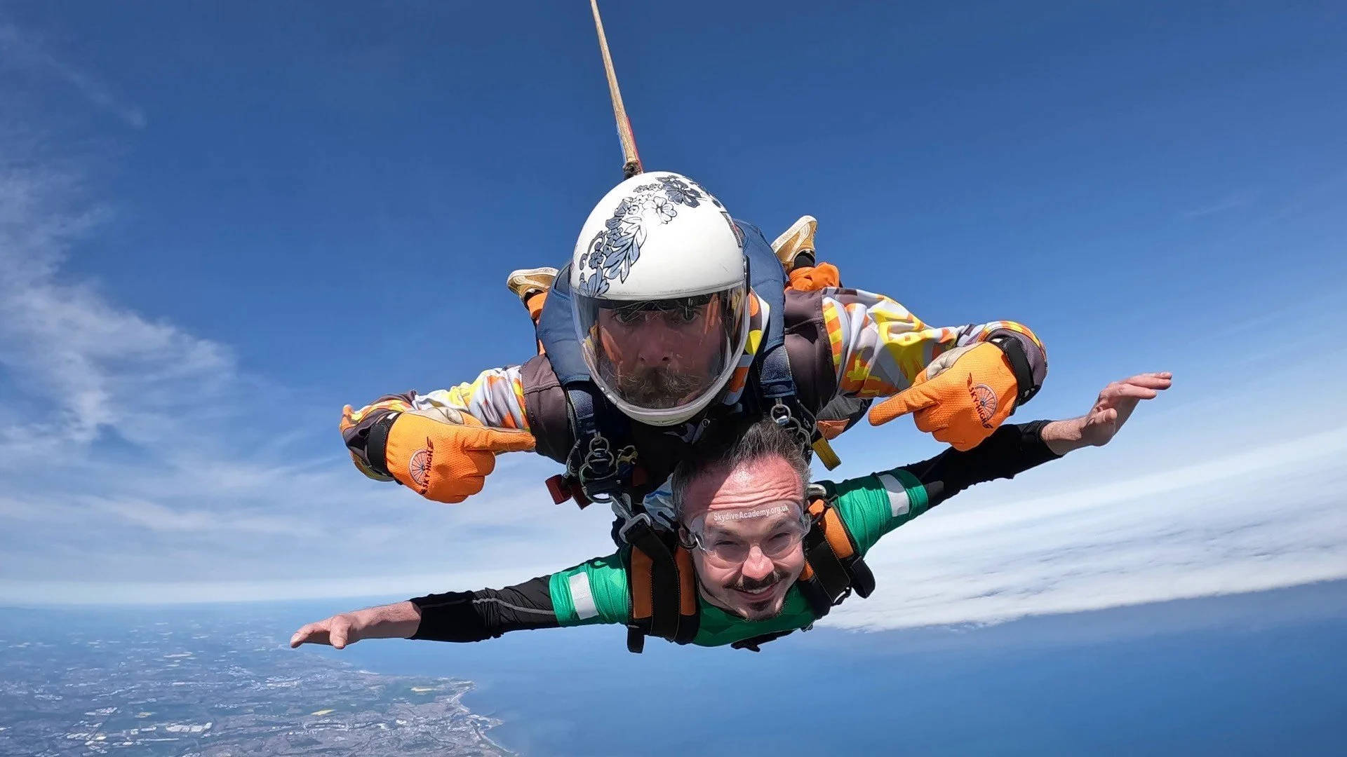 Two skydivers free-falling high above the coastline, one in a helmet and goggles holding onto the other, with the ocean and land visible below and a clear blue sky.