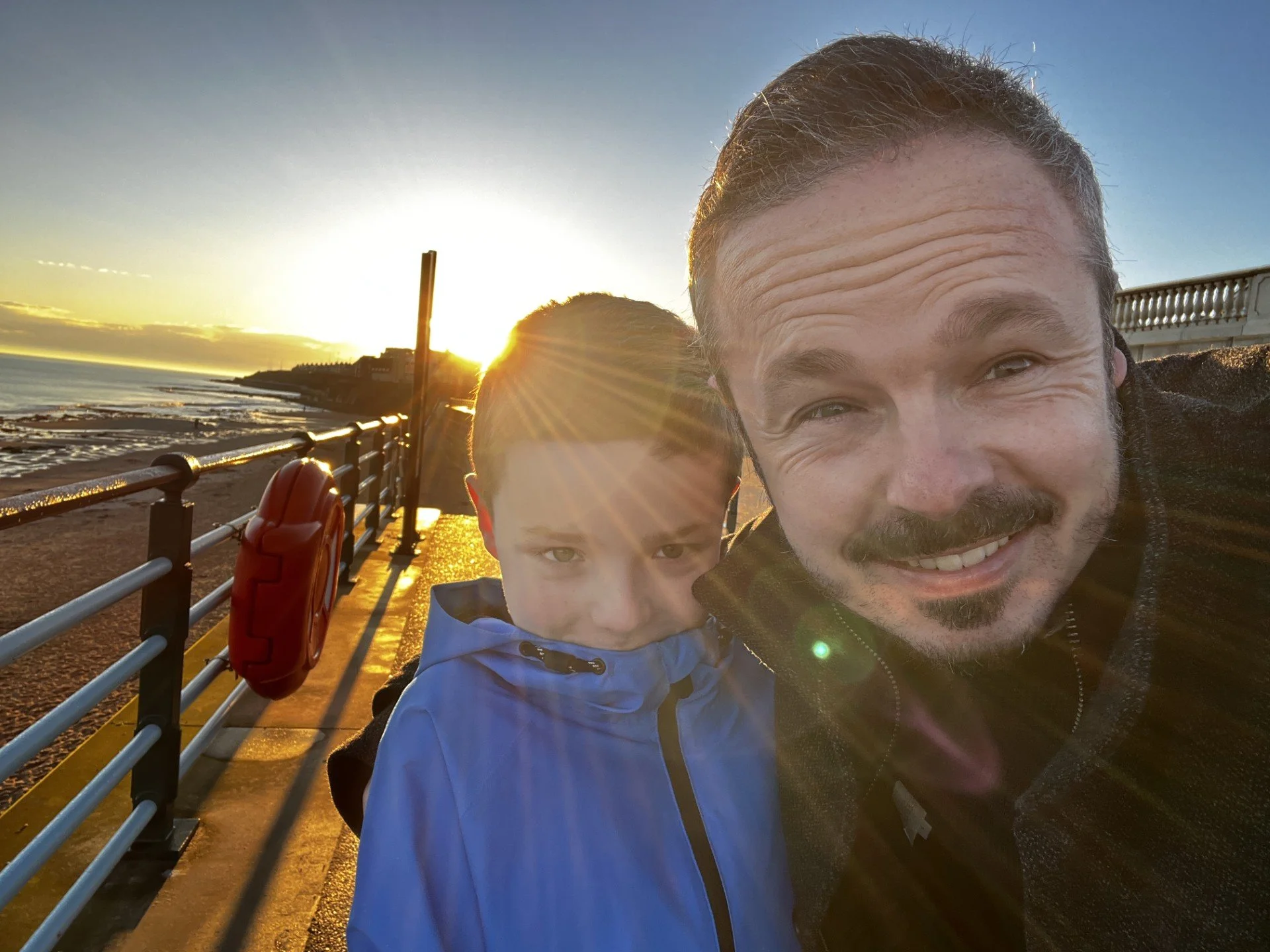 A man and a boy taking a selfie on a pier at sunset, with the ocean and a hill in the background.