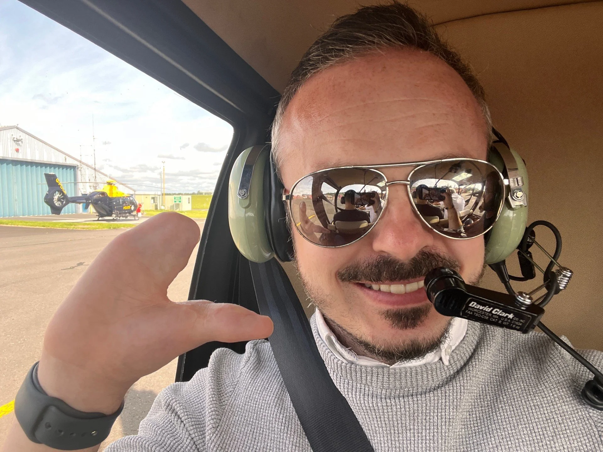 Smiling man in sunglasses and headset inside a helicopter taking a selfie, with police helicopter visible outside on tarmac.