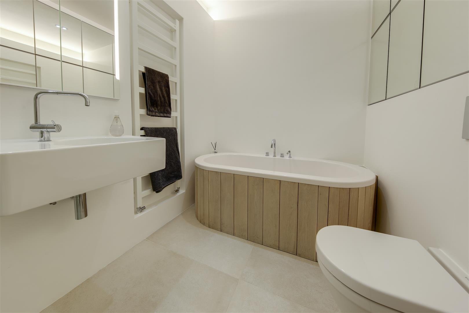 Modern bathroom with a wall-mounted sink, a freestanding bathtub with a wooden side panel, a toilet, and mirrored cabinets.