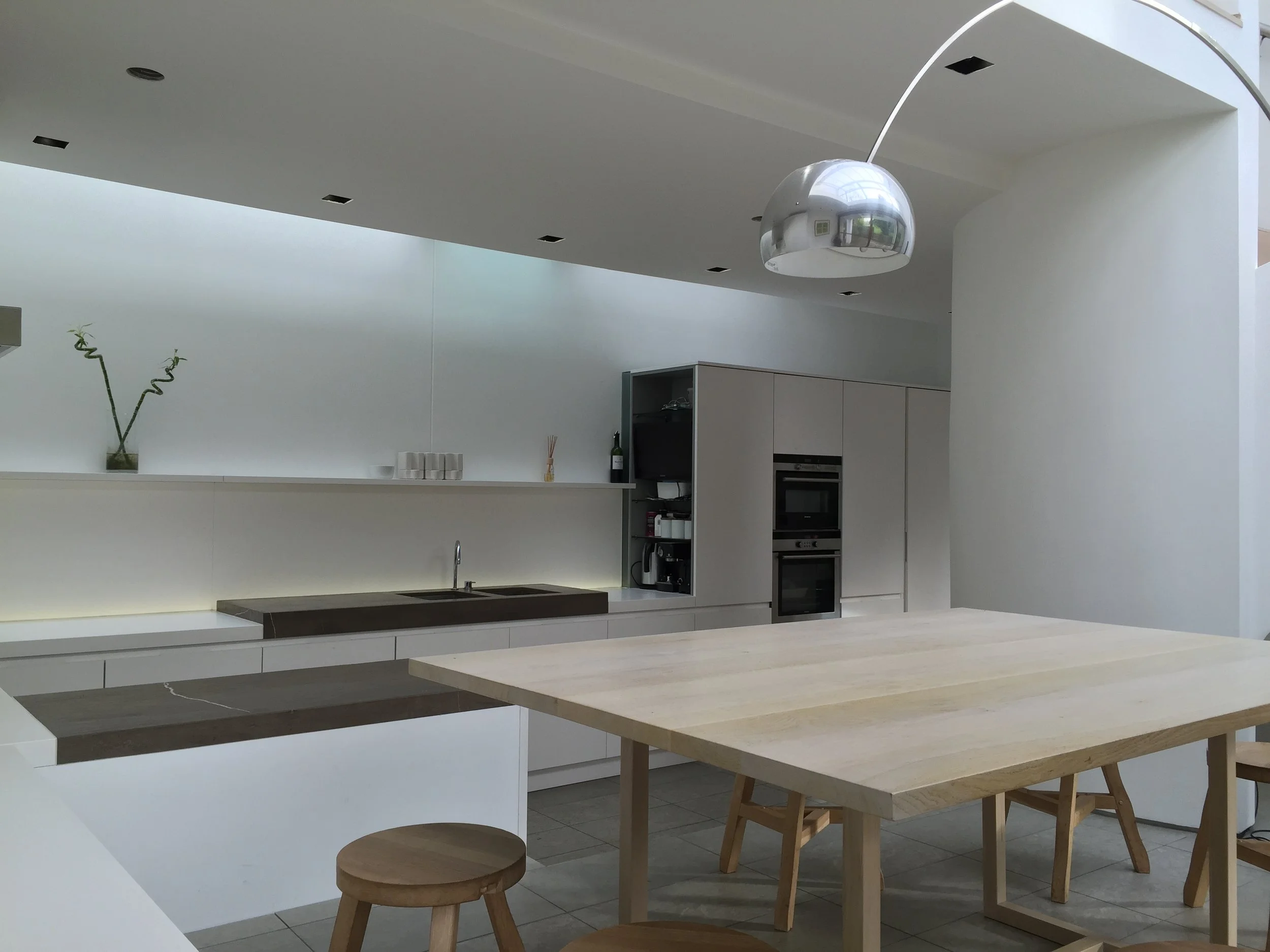 Modern minimalist kitchen with white cabinets, a wooden dining table, stools, built-in ovens, and a metallic pendant lamp.