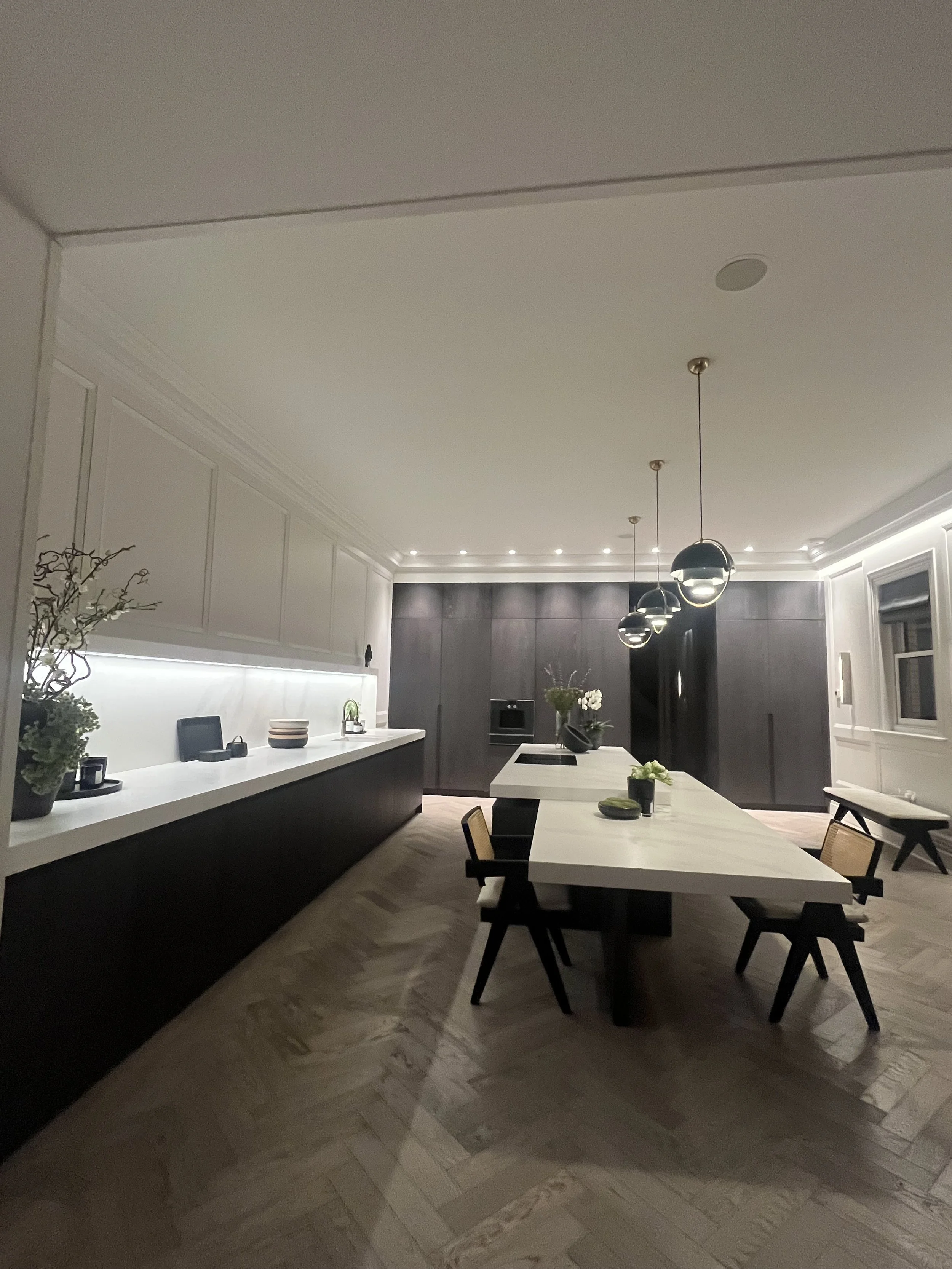 Modern kitchen with a long white island, black stools, pendant lights, and dark cabinetry at the far end, decorated with plants and bowls.