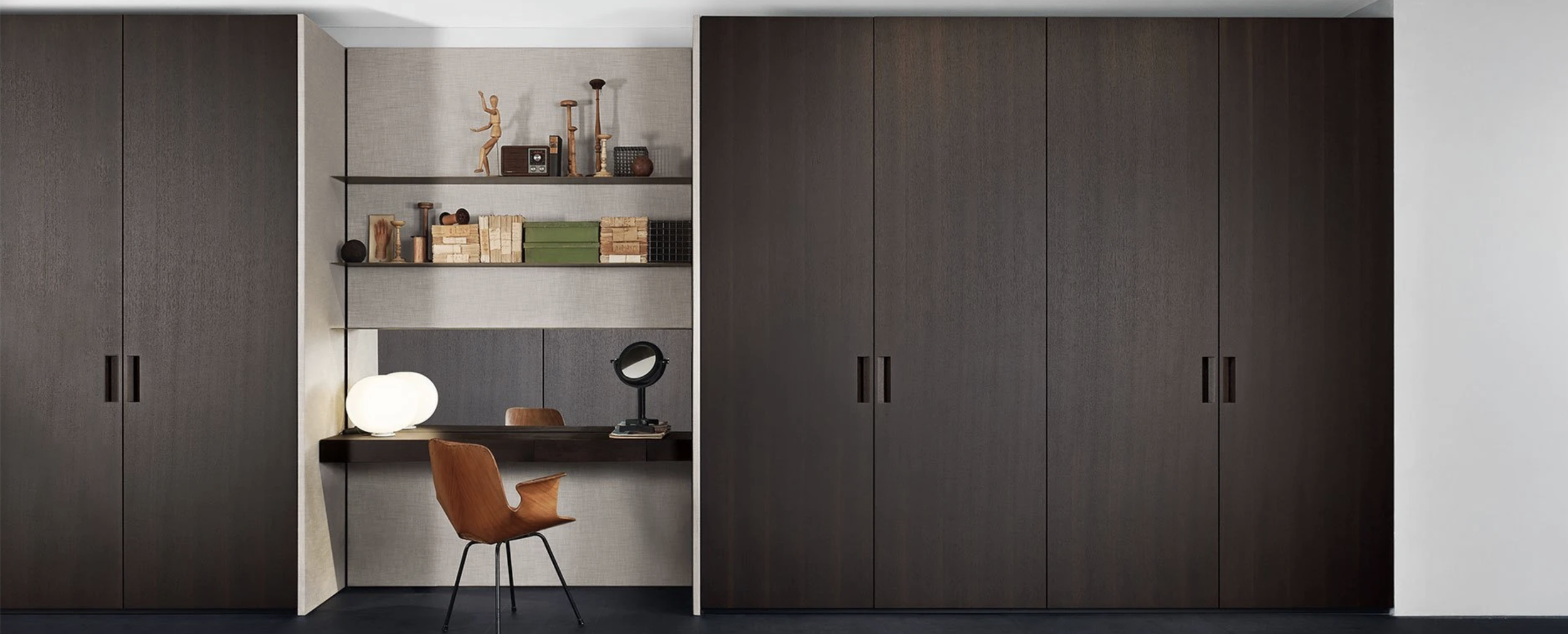 A modern built-in study nook with dark wood cabinets, open shelving with decorative items, and a desk with a lamp, mirror, and a wooden chair.