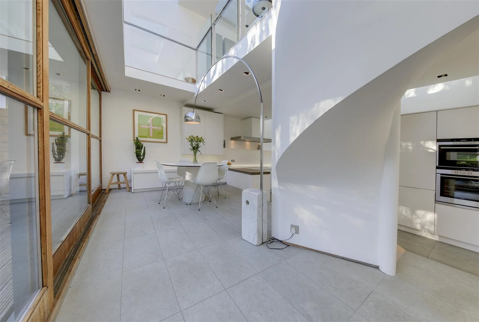 Modern kitchen and dining area with white walls, a white dining table with white chairs, indoor plants, and large glass windows and sliding doors.