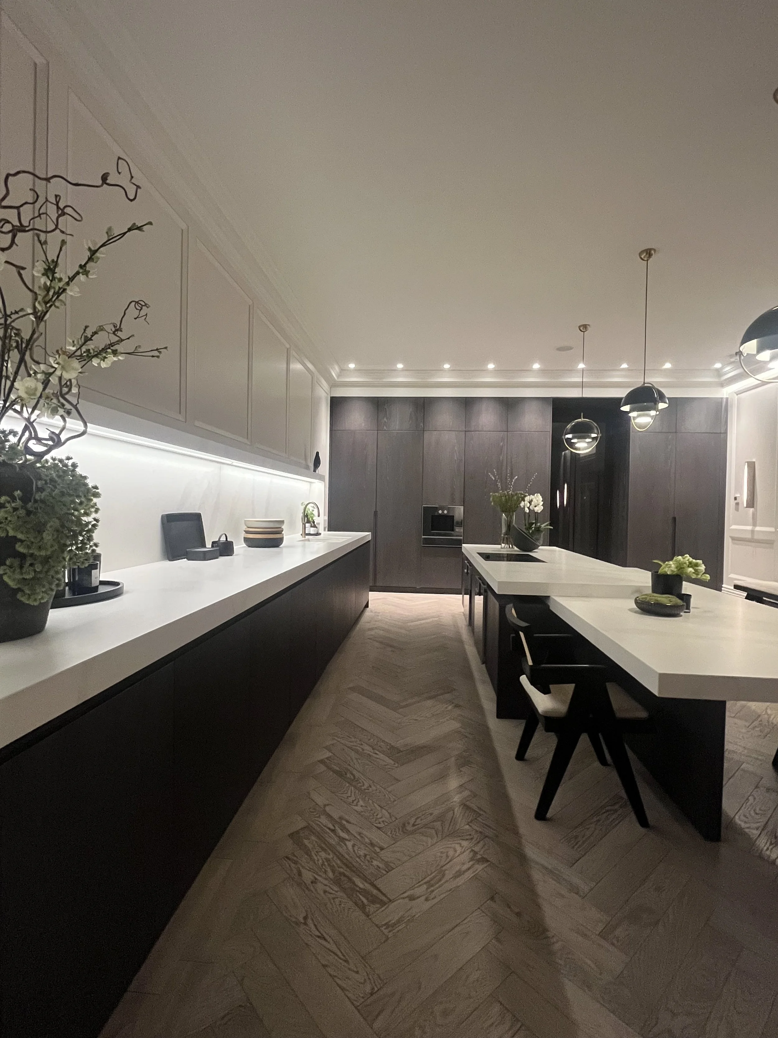 Modern kitchen with black and white cabinetry, a long white countertop, pendant lighting, and decorative plants.