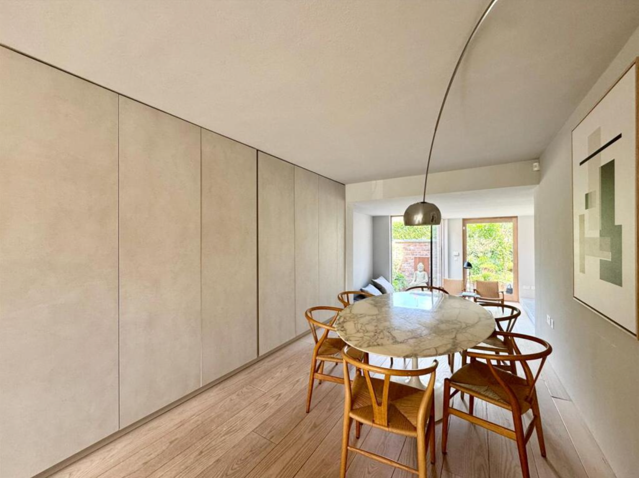 Dining area with a marble round table and six wooden chairs, large windows with outdoor greenery, light-colored walls, and modern decor.