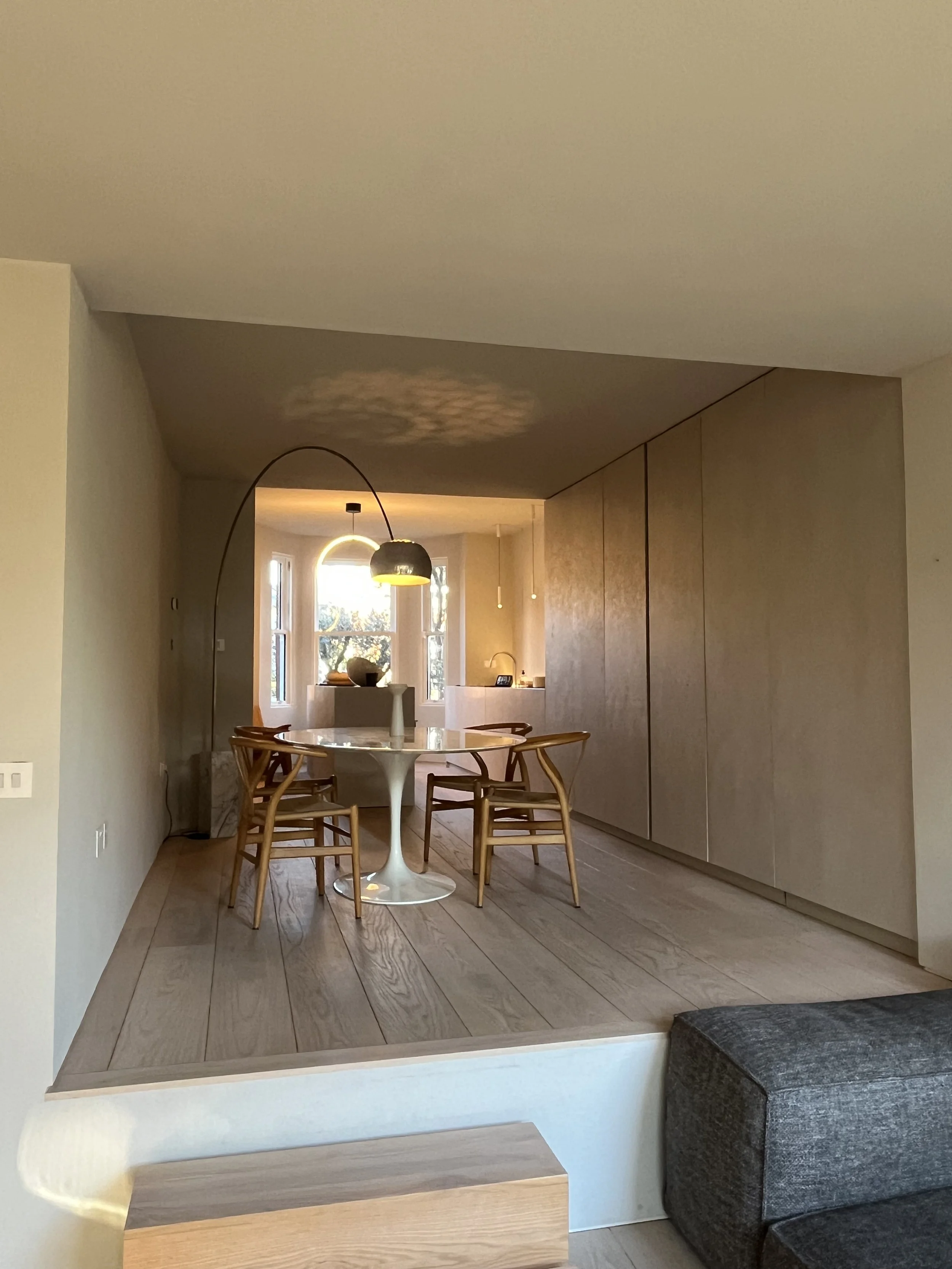 A dining area with a round white table, four wooden chairs with curved backs, a modern arc floor lamp with a black shade, and a large window letting in natural light; adjacent to a gray upholstered sofa.