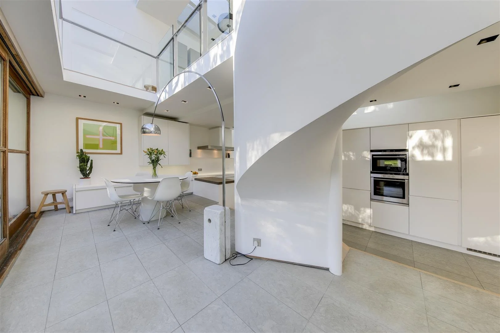 Modern, bright kitchen and dining area with white walls, tiled floor, and large sliding glass door. Balcony or loft area with glass railing visible upstairs. Dining table with white chairs, plants on a sideboard, and built-in white cabinets with doub