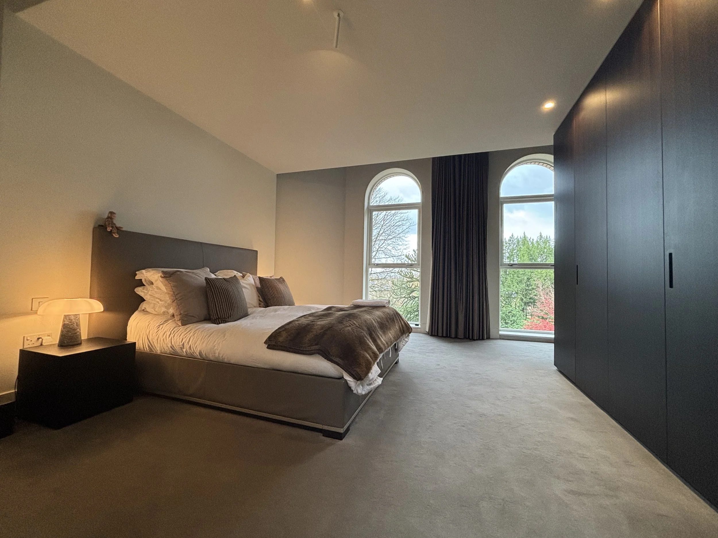 A modern bedroom with a large bed featuring gray and white bedding and pillows, a black nightstand with a lamp, two arched windows with dark curtains, and a dark built-in wardrobe to the right.