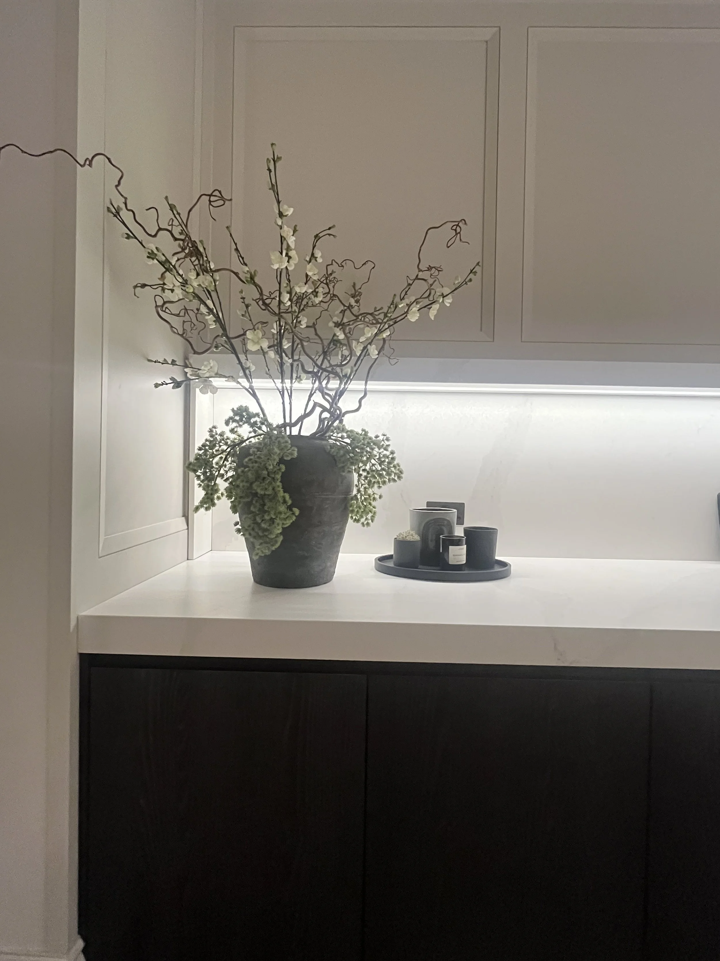 A decorative indoor arrangement with a black vase containing curly willow branches and green and white flowers on a white countertop, with small black candles on a tray nearby.