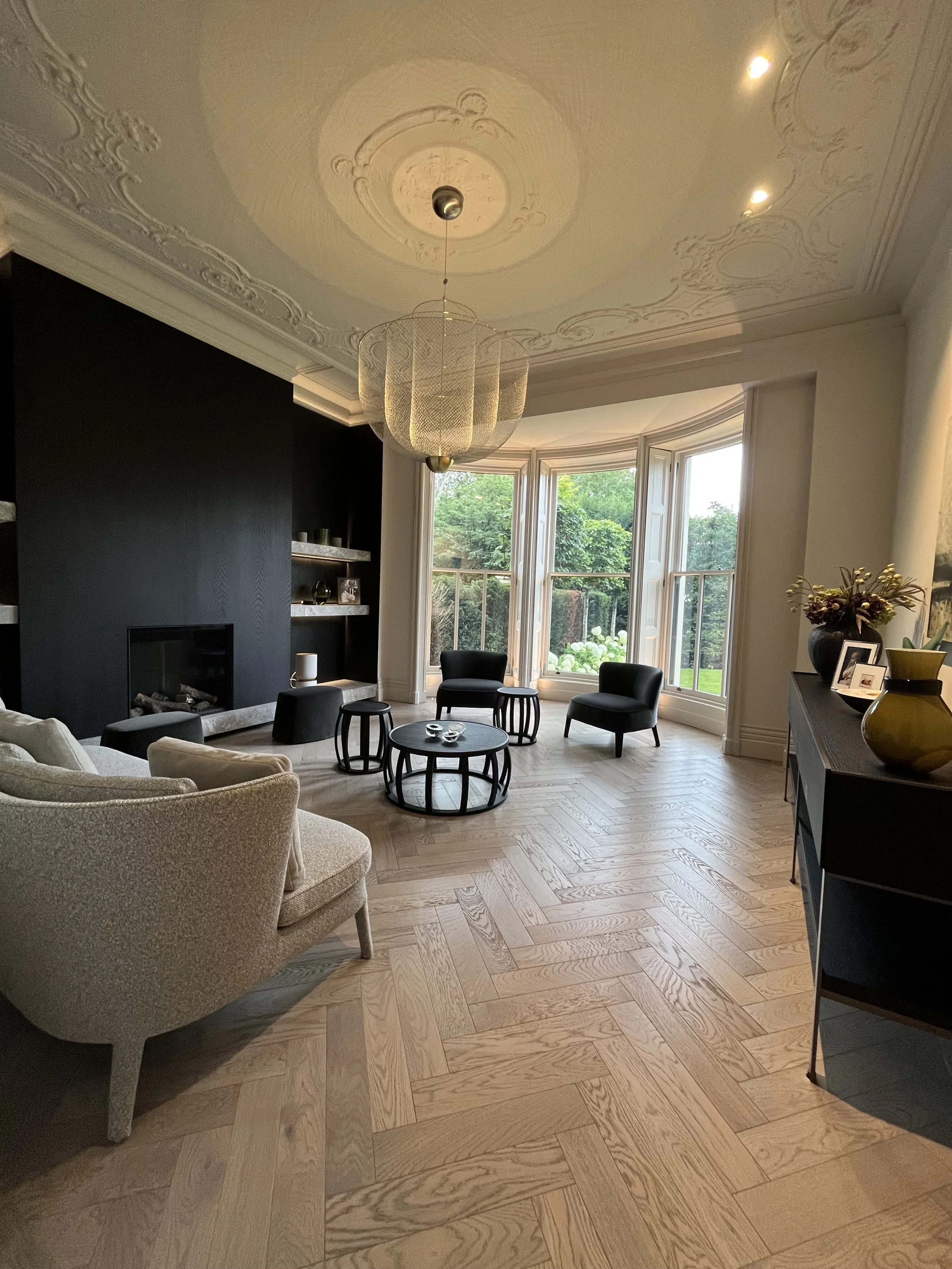 Living room with a beige armchair, black chairs, a black sideboard, large bay window, wooden herringbone floor, ornate ceiling, decorative chandelier, and view of greenery outside.