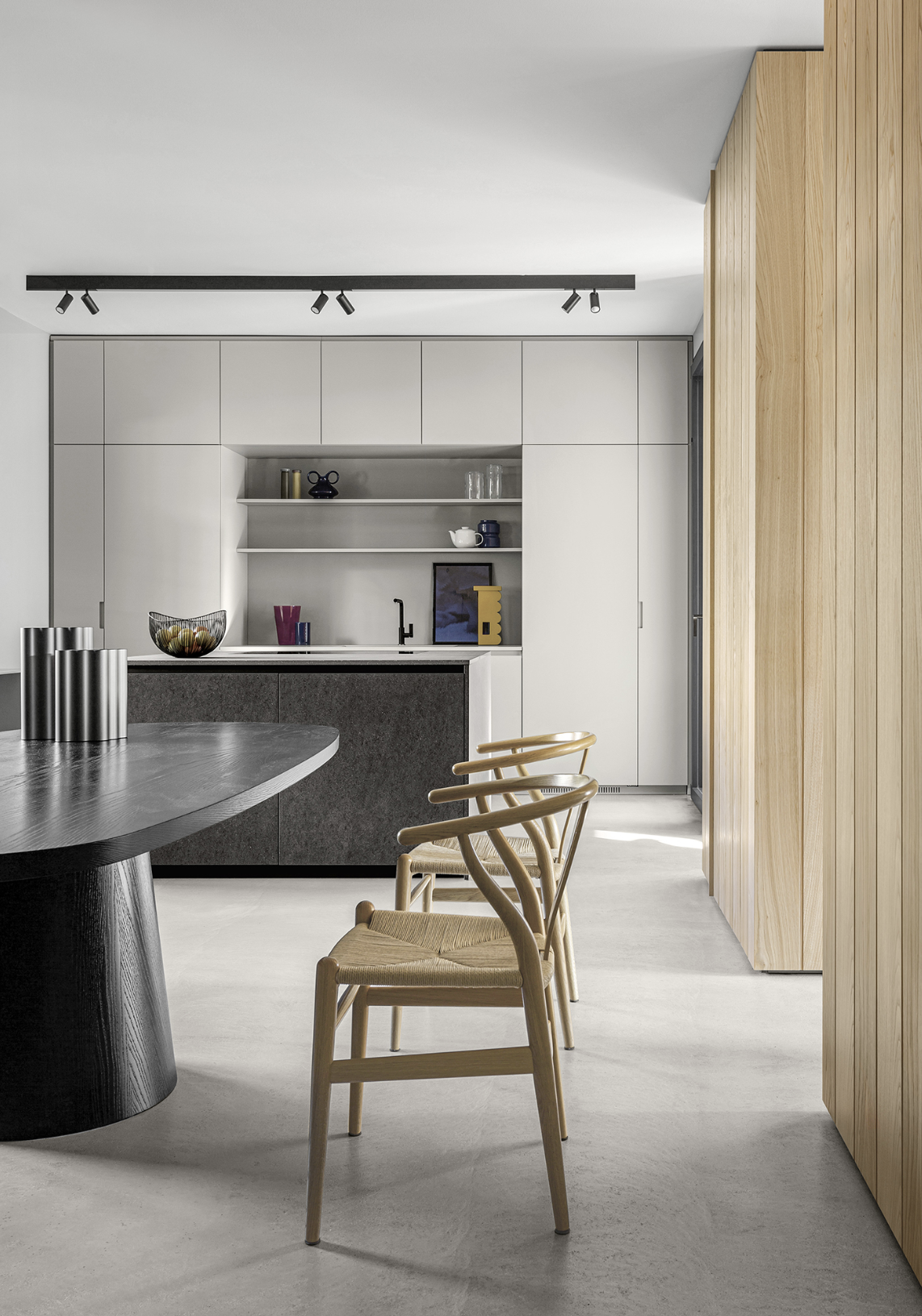 Modern kitchen with light wooden cabinets, a black island, white walls, and a concrete floor. There are three rattan chairs around a dark round table, and open shelves with decorative items on the wall behind the sink.