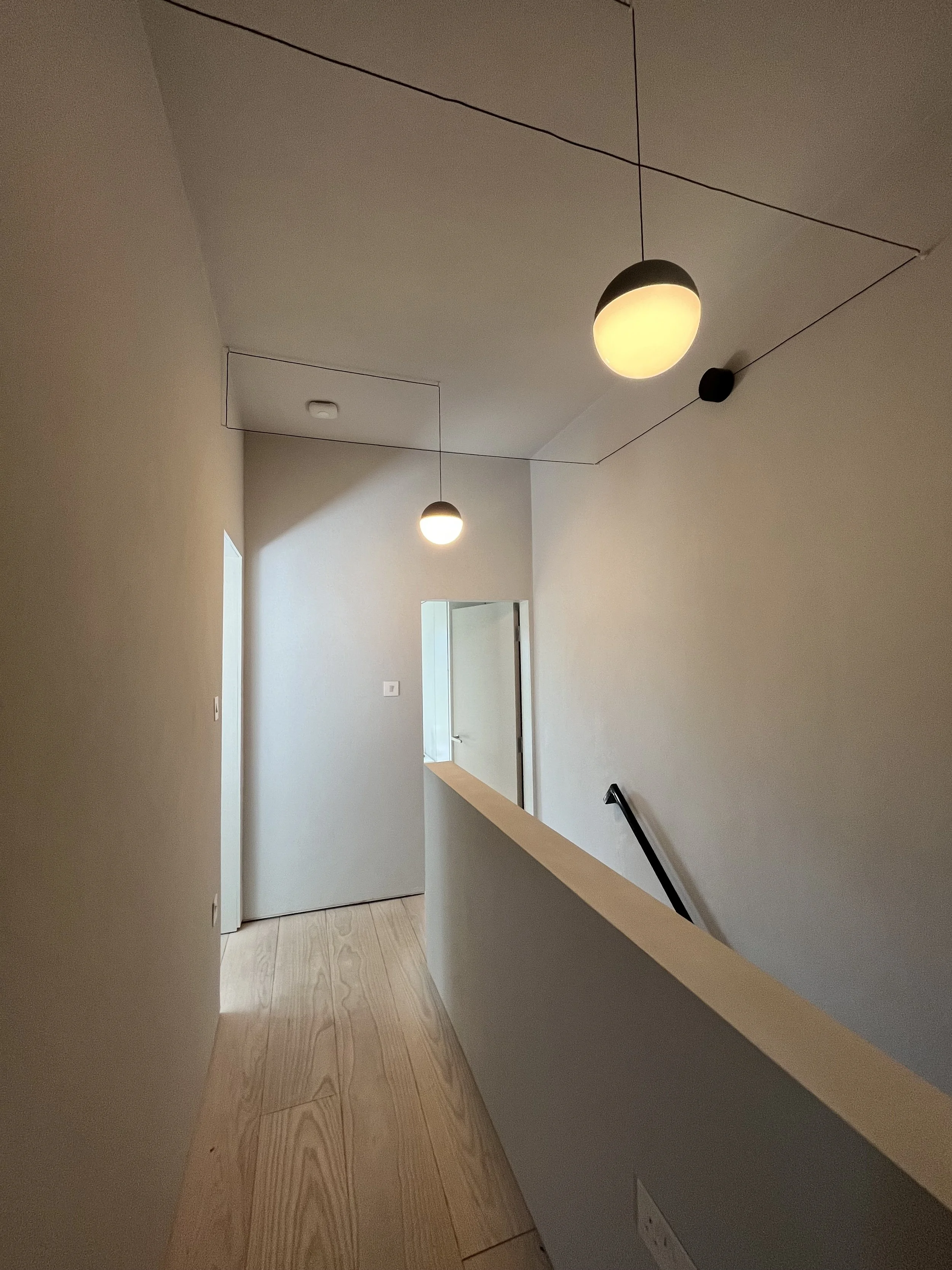 A hallway with light-colored walls and wooden flooring, illuminated by two ceiling lights, with a black handrail on the right side and doors at the end.