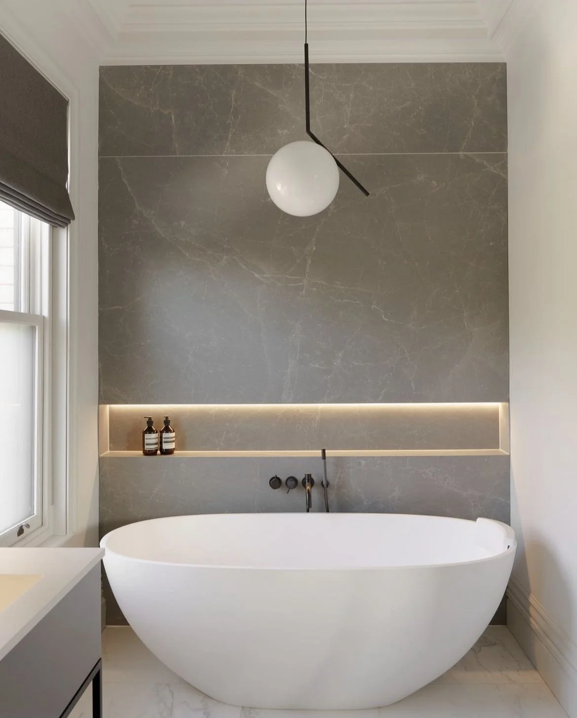 Modern bathroom with round white bathtub, gray marble wall, built-in shelf with two bottles, black faucet, pendant light, and window with horizontal blind.