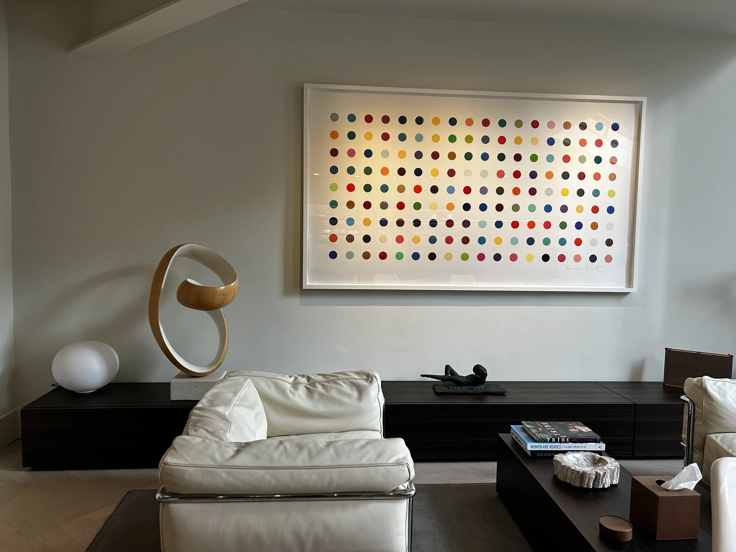 Modern living room with white leather sofa, black coffee table holding books and decorative object, dark wood sideboard with artwork and sculptures, and framed colorful polka dot artwork on light gray wall.