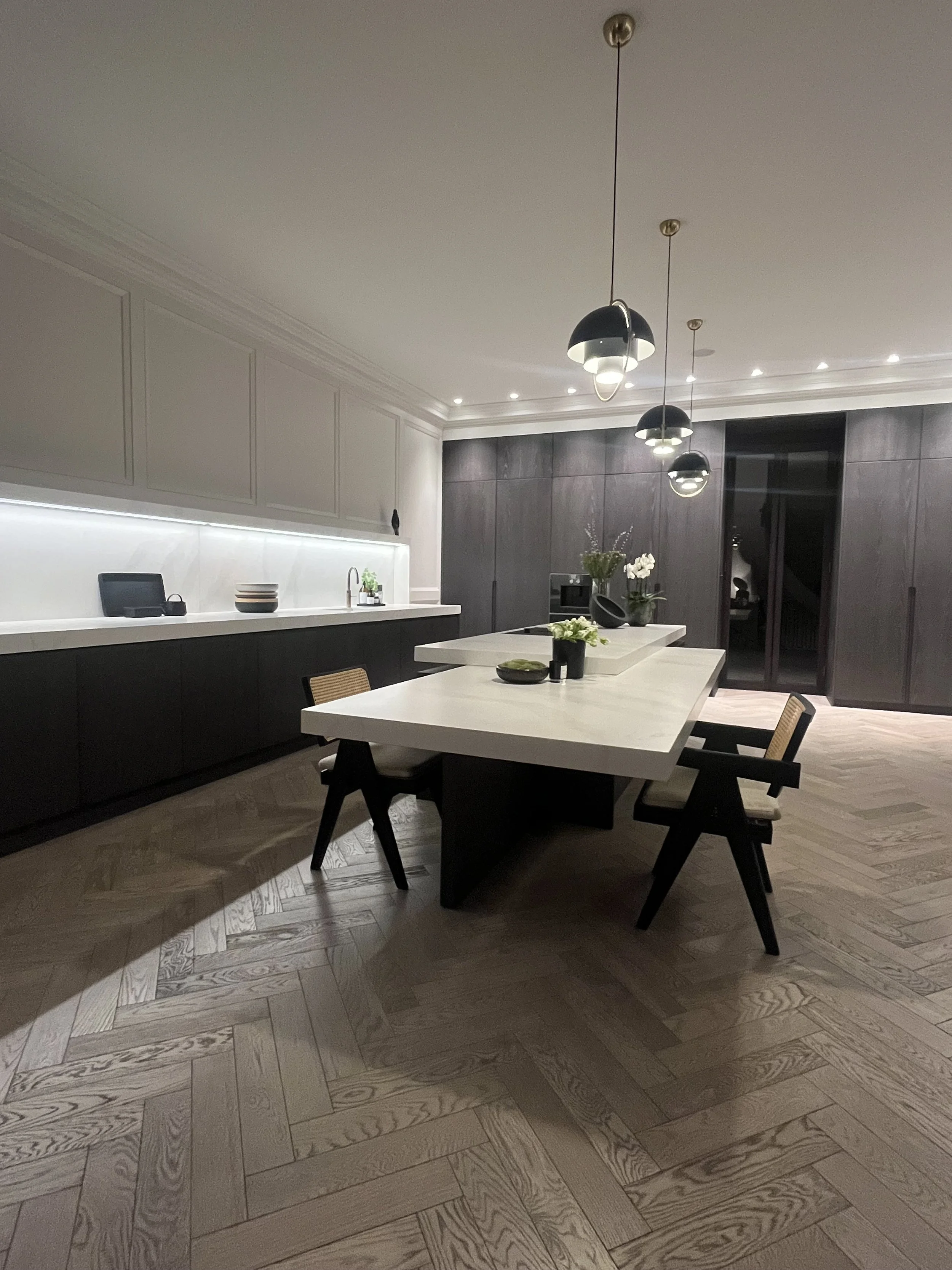 Modern kitchen and dining area with a large white table, black chairs, dark wood cabinetry, pendant lighting, and decorative plants.