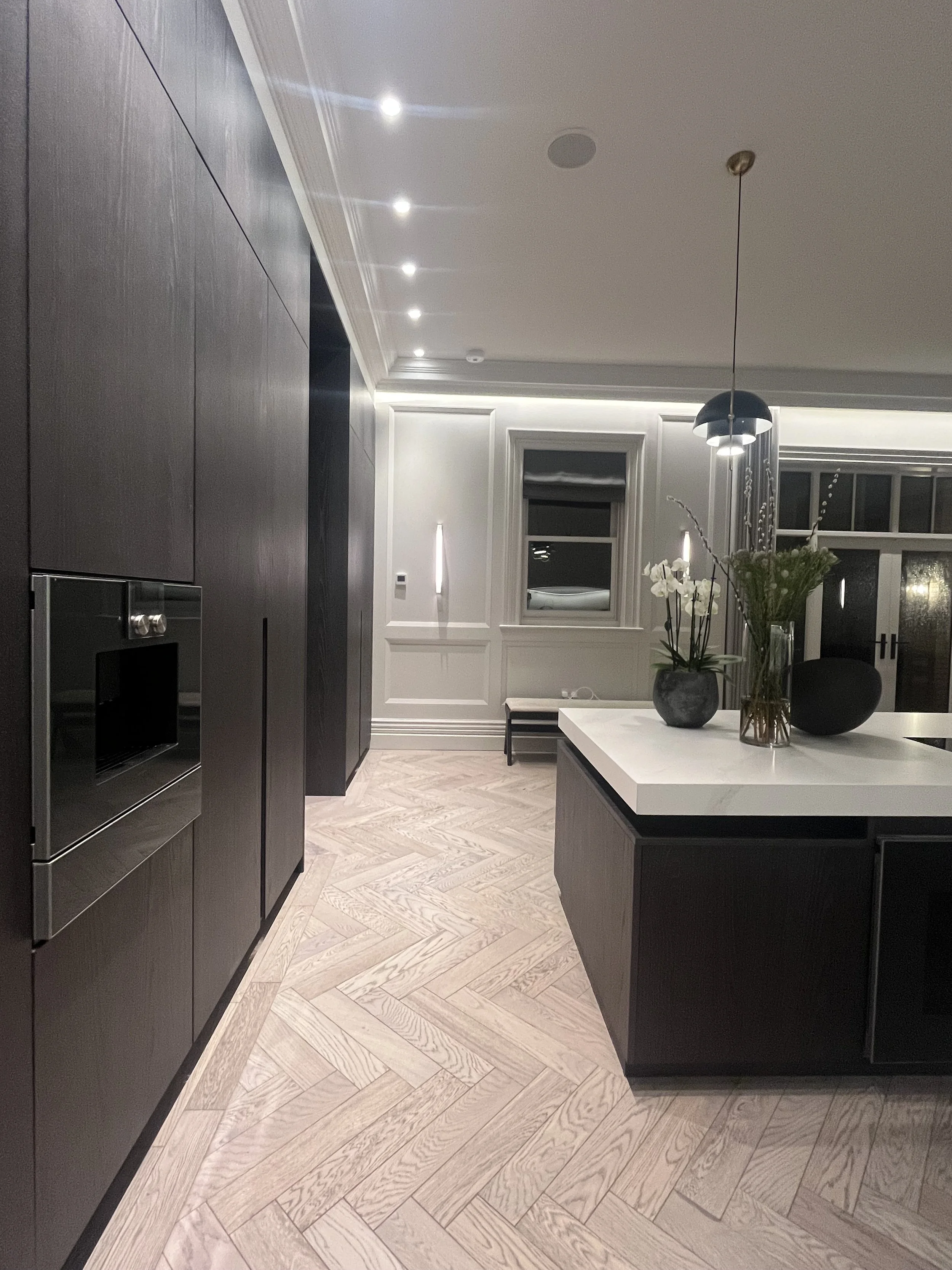 Modern kitchen with dark cabinets, white countertop, herringbone patterned light wood floor, pendant light, and decorative vases with flowers on the island.
