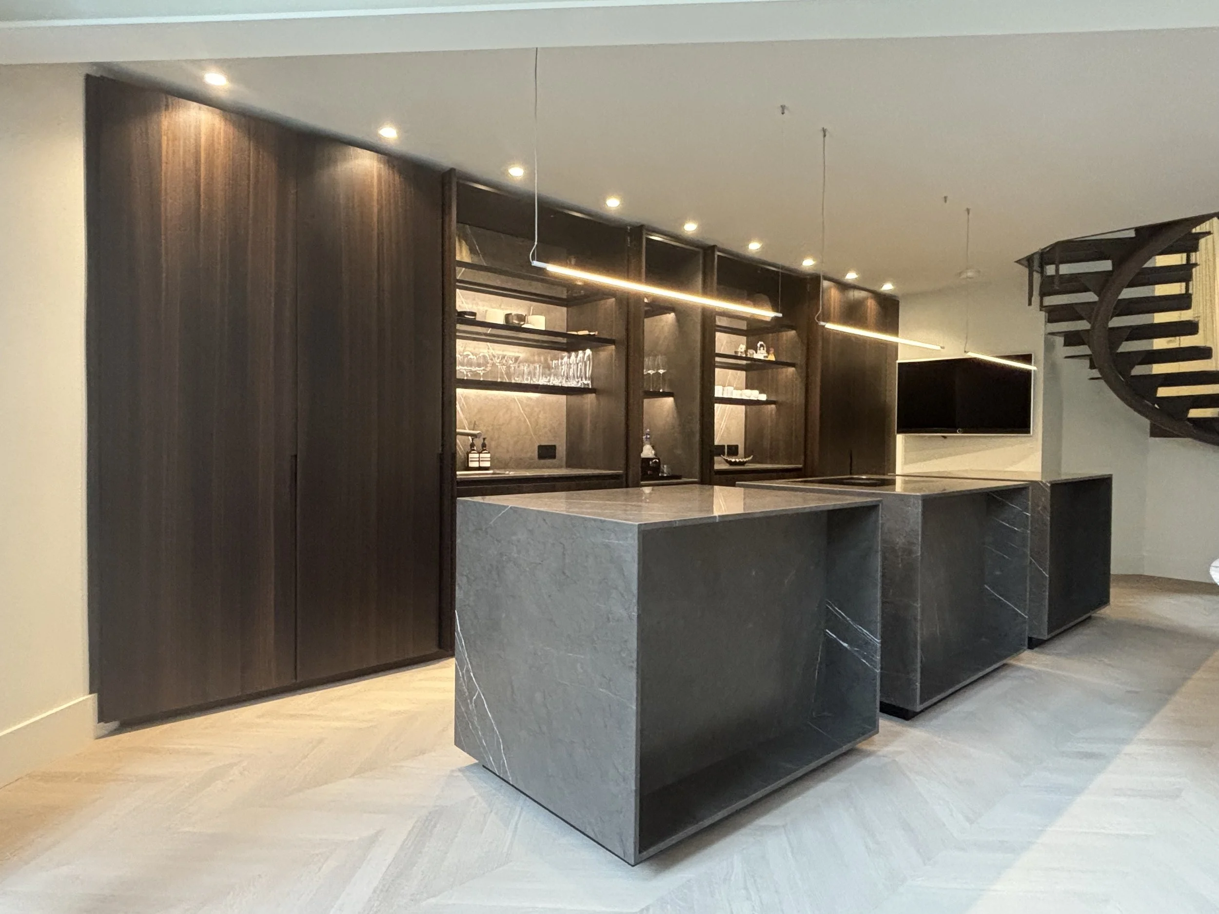 Modern kitchen with dark wood cabinets, marble countertops, open shelving, and a mounted flat-screen TV, near a spiral staircase.