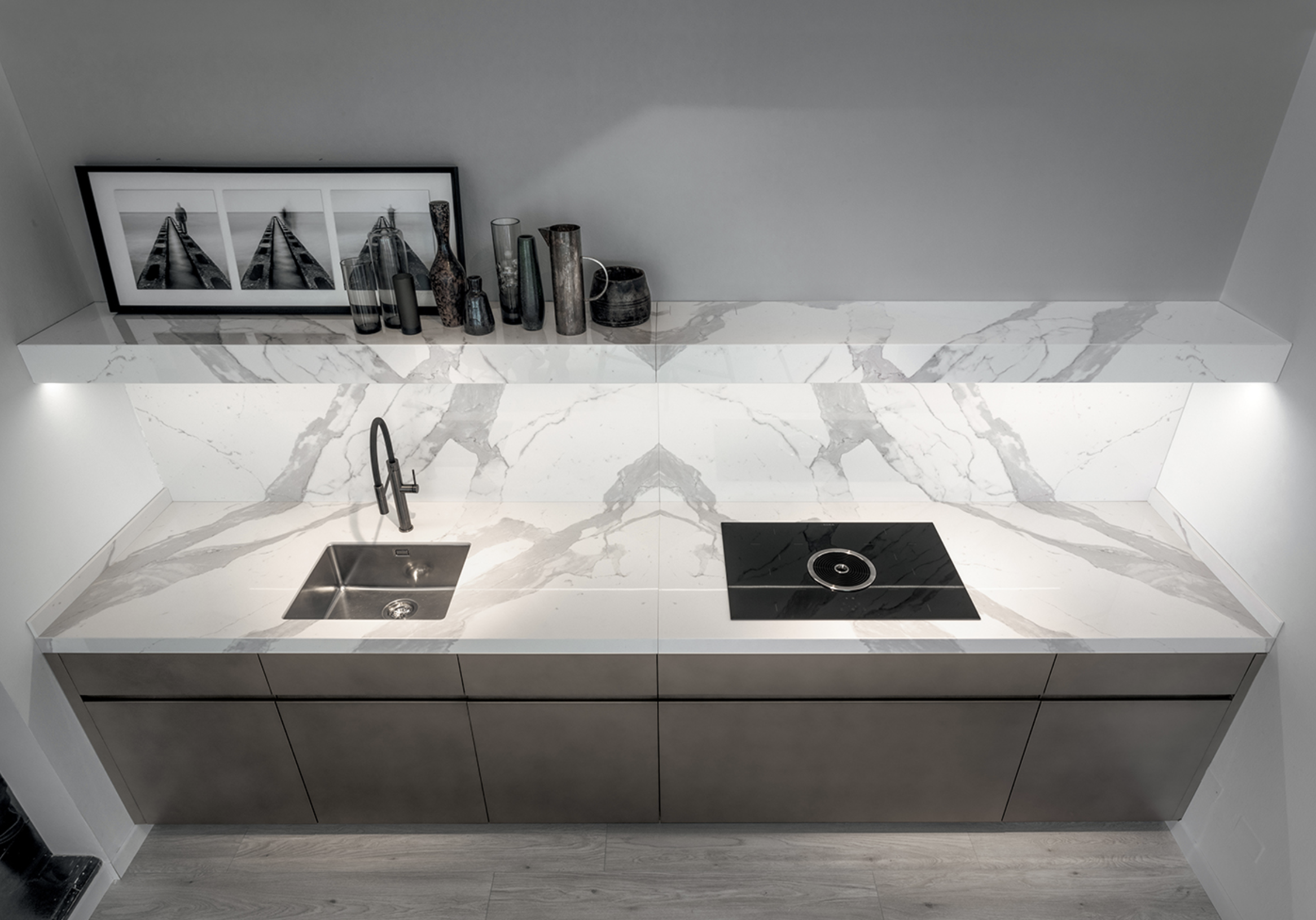 Modern kitchen countertop with white marble surface, stainless steel sink, black electric cooktop, grey cabinets, and decorative vases and framed art on a shelf in the background.