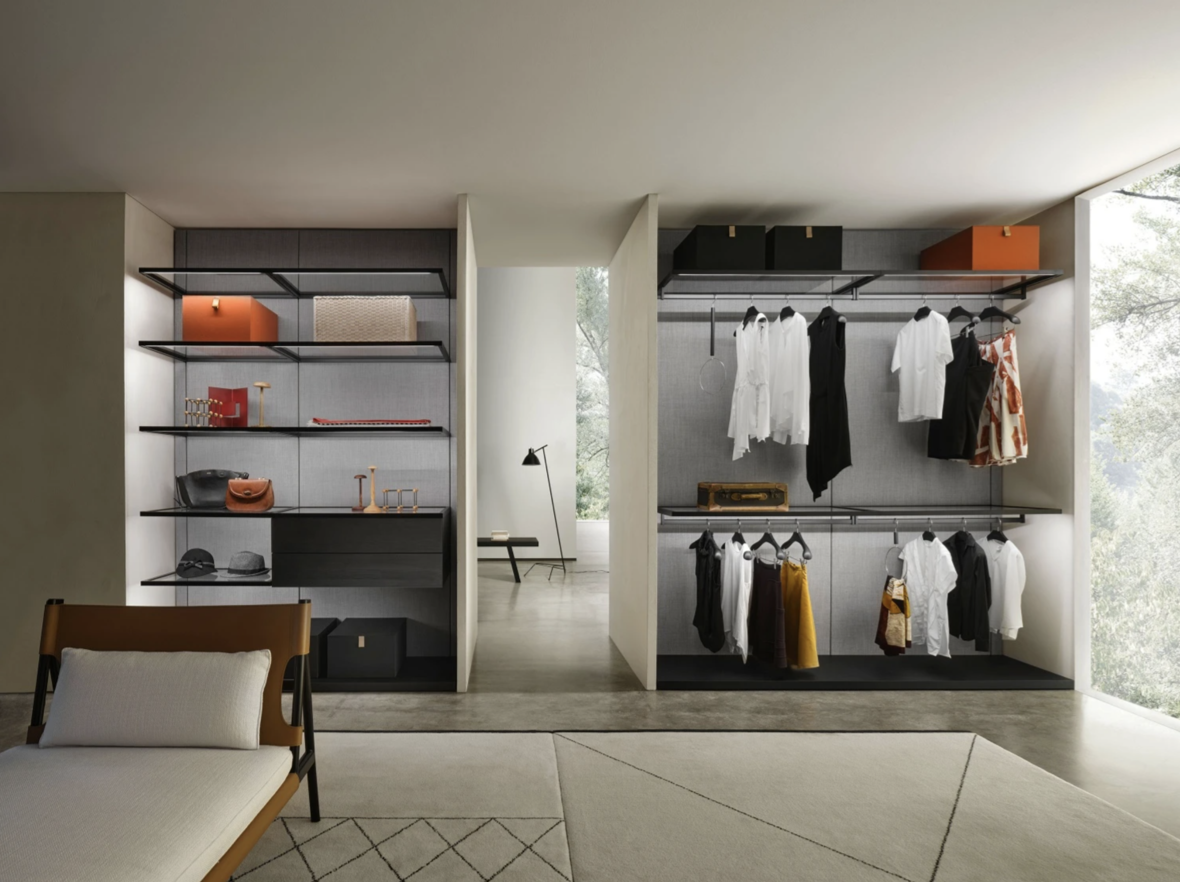 Modern walk-in closet with black shelving, hanging clothes, and storage boxes, next to a seating area with a chair and a patterned rug, illuminated by natural light from large windows.