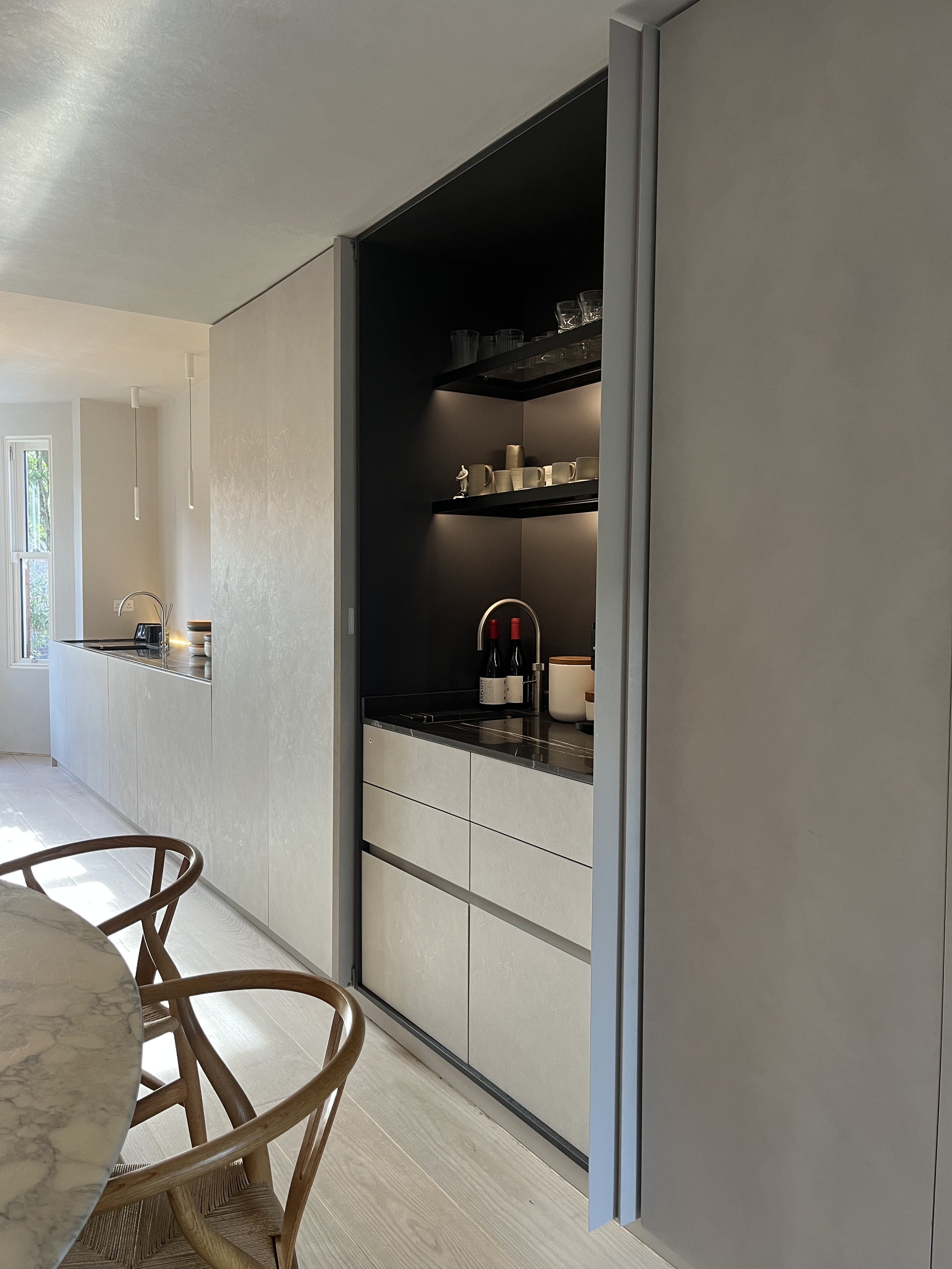 Modern kitchen with built-in cabinetry, a black backsplash, wine bottles, cups, glasses, and a marble dining table with wooden chairs.