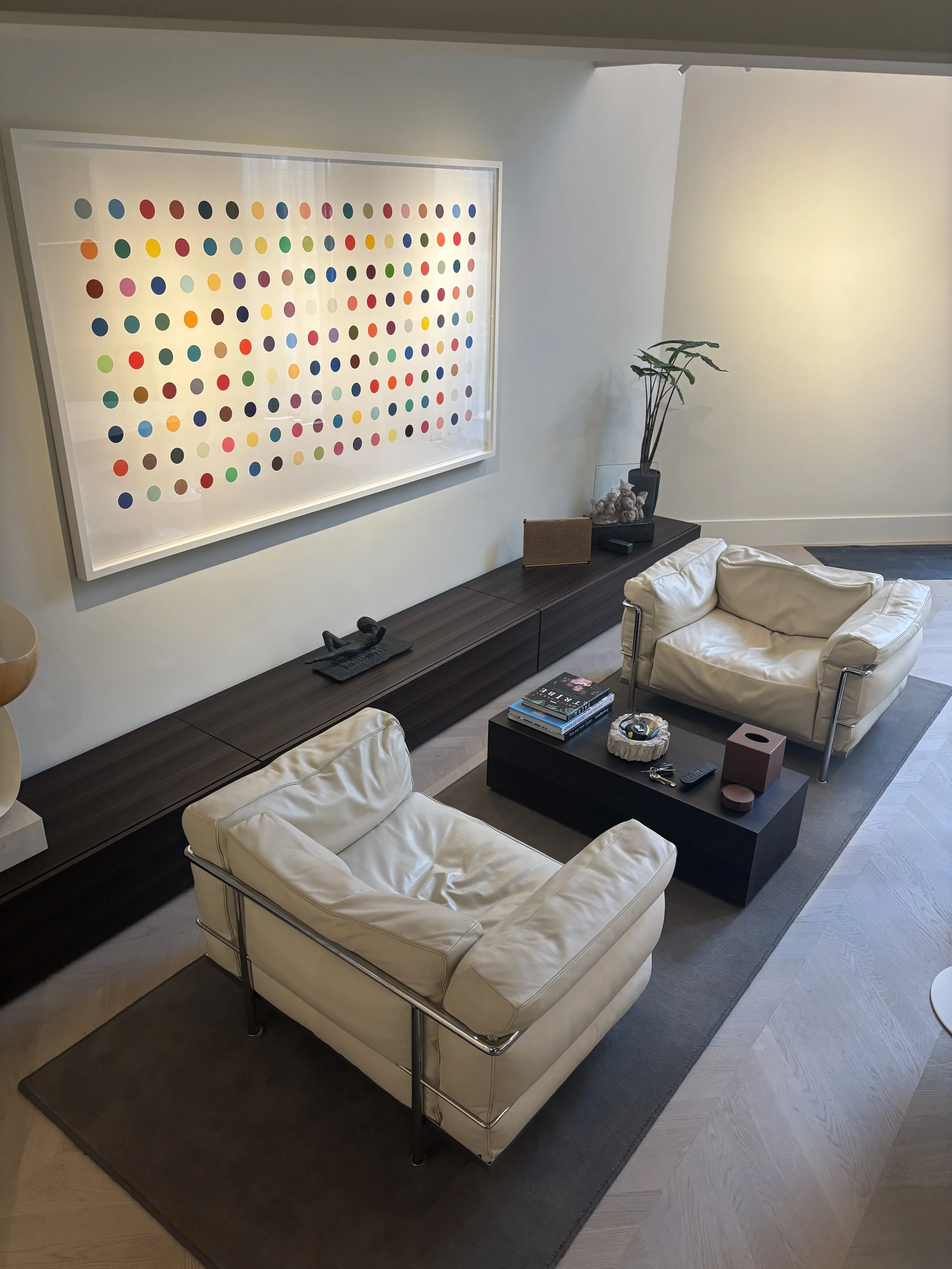 Modern living room with two white leather armchairs, a low black coffee table with books, a decorative box, keys, and a remote. A long dark wooden console runs along the wall with a sculpture, a wooden box, and a tall plant. Above the console is a la