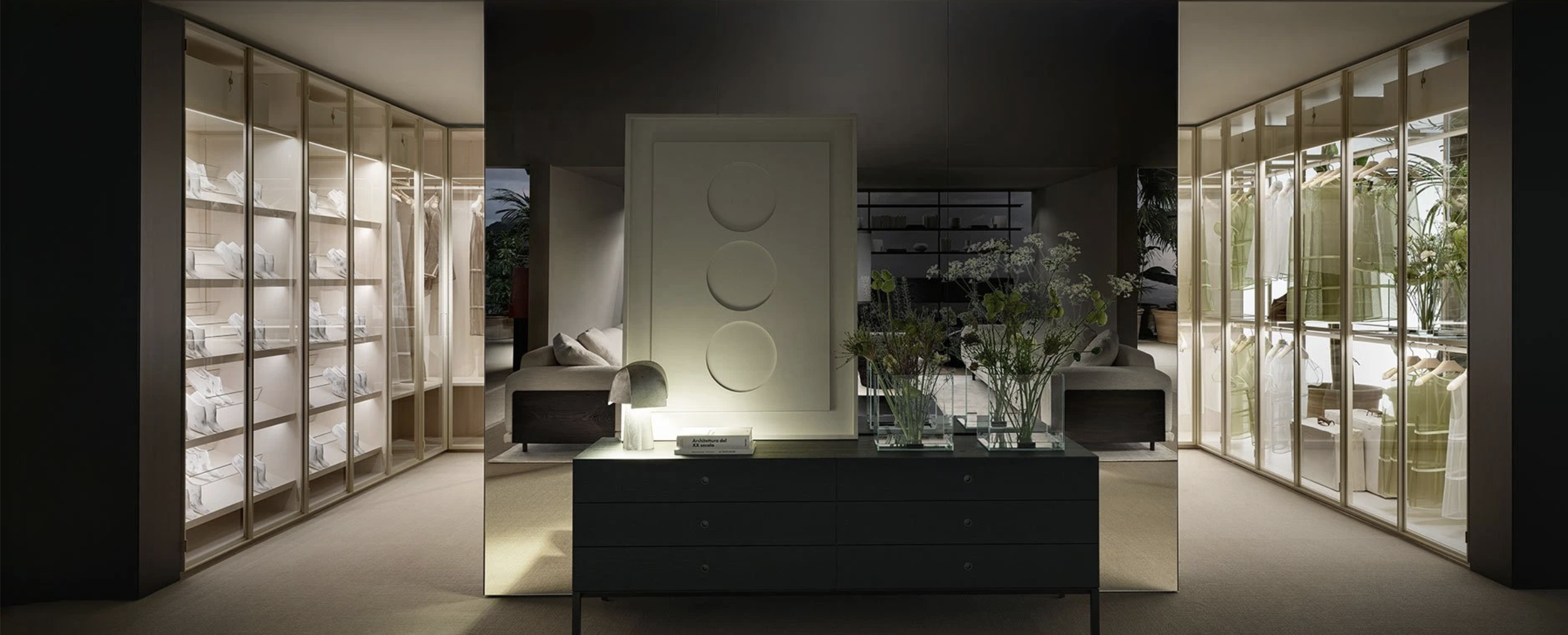 Modern walk-in closet with glass-fronted wardrobe units displaying clothing, and a dark dresser with vases of flowers in the center.
