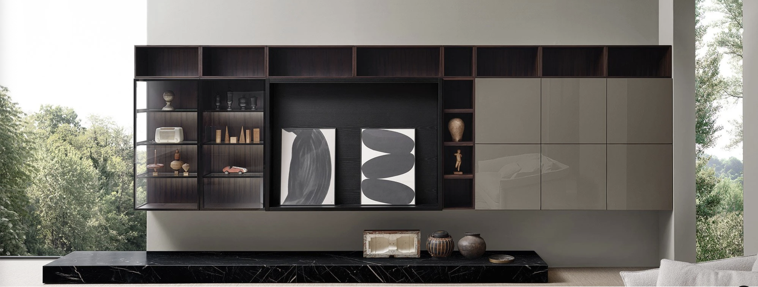 Modern wall unit with open and closed storage, decorated with artwork, vases, and small sculptures, in a room with large windows showing green trees outside.
