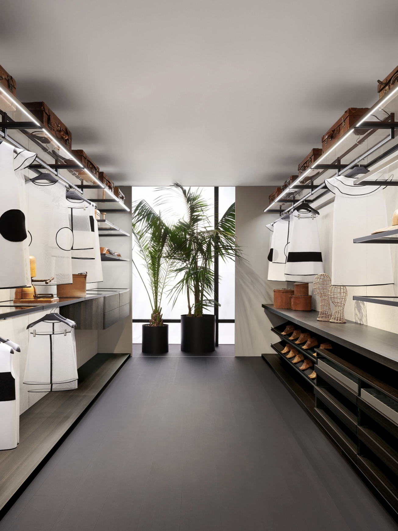 Empty boutique with clothing on display, large potted plants at back, minimalist design with neutral colors and modern lighting.