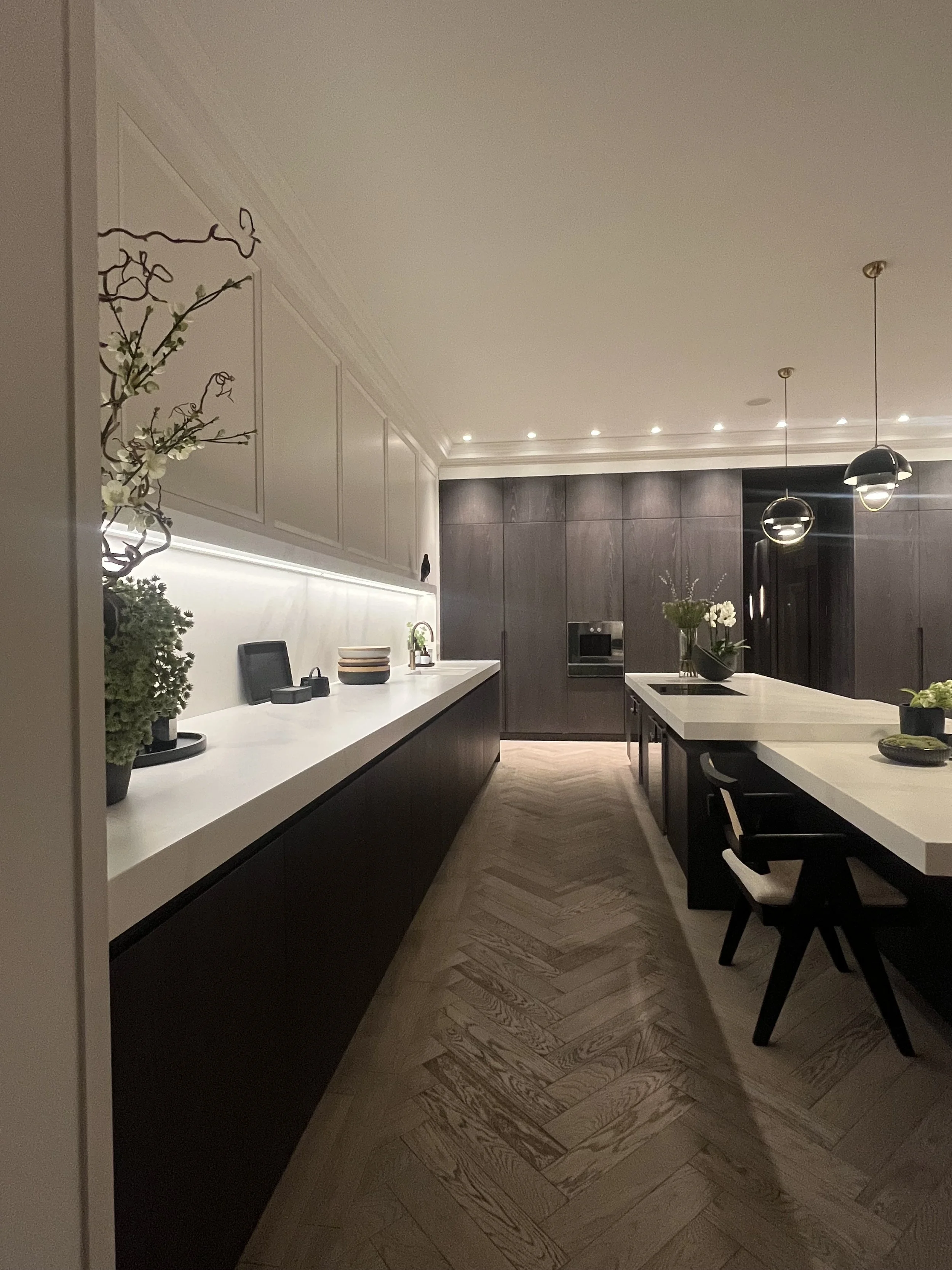 Modern kitchen with white countertops, dark cabinets, and decorative plants, illuminated by ceiling lights and pendant lighting.