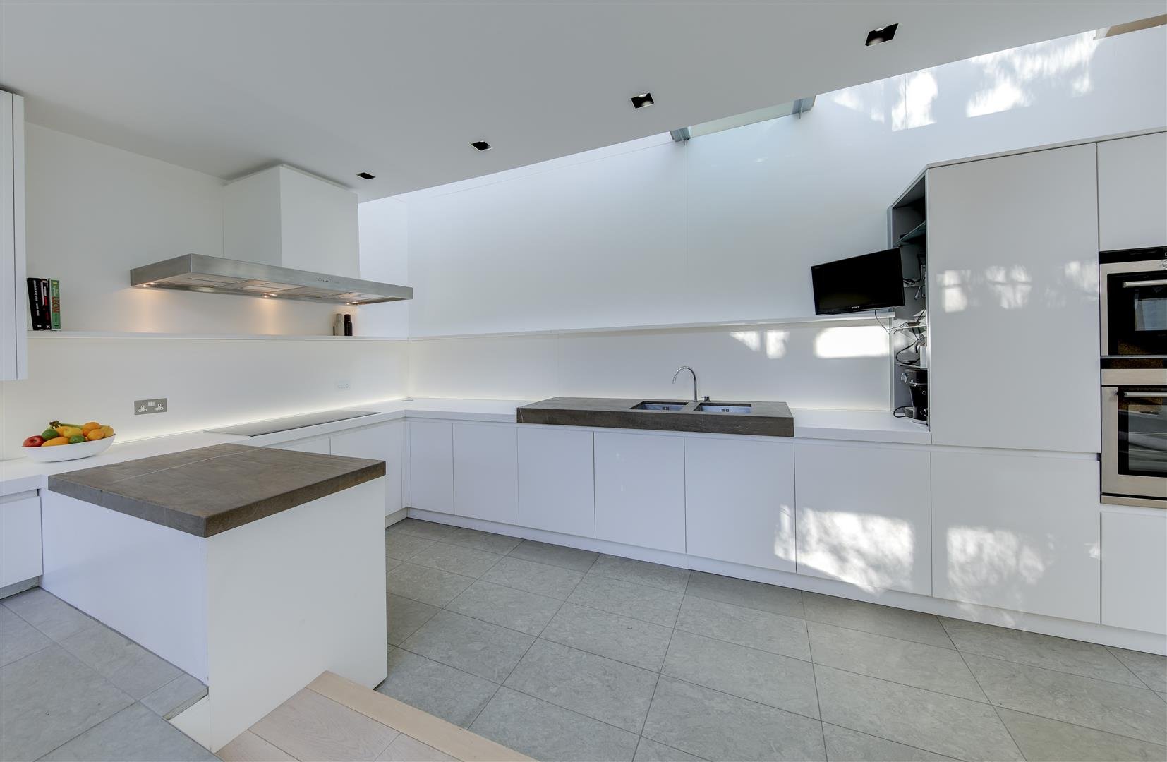 Modern kitchen with white cabinets, a gray countertop island, a stainless steel range hood, and a built-in oven, featuring natural light and minimalistic design.
