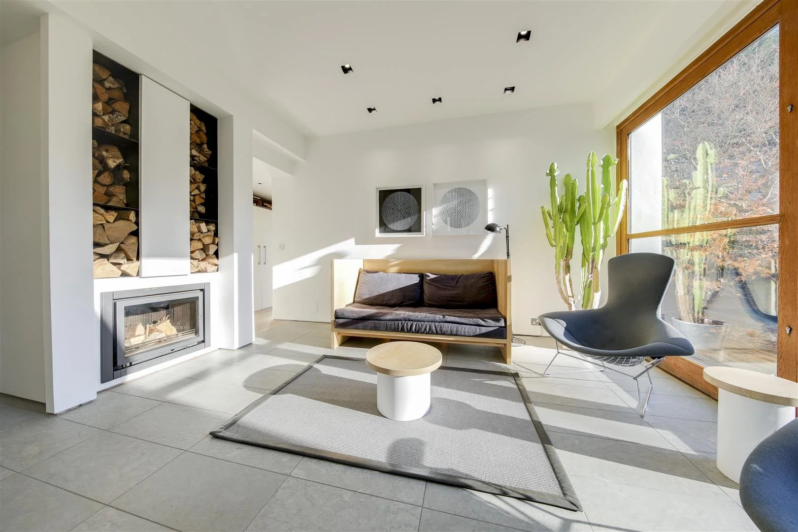 Minimalist living room with wood-burning fireplace, stacked firewood, modern black lounge chair, small round coffee table, large window with cacti outside, and abstract wall art.