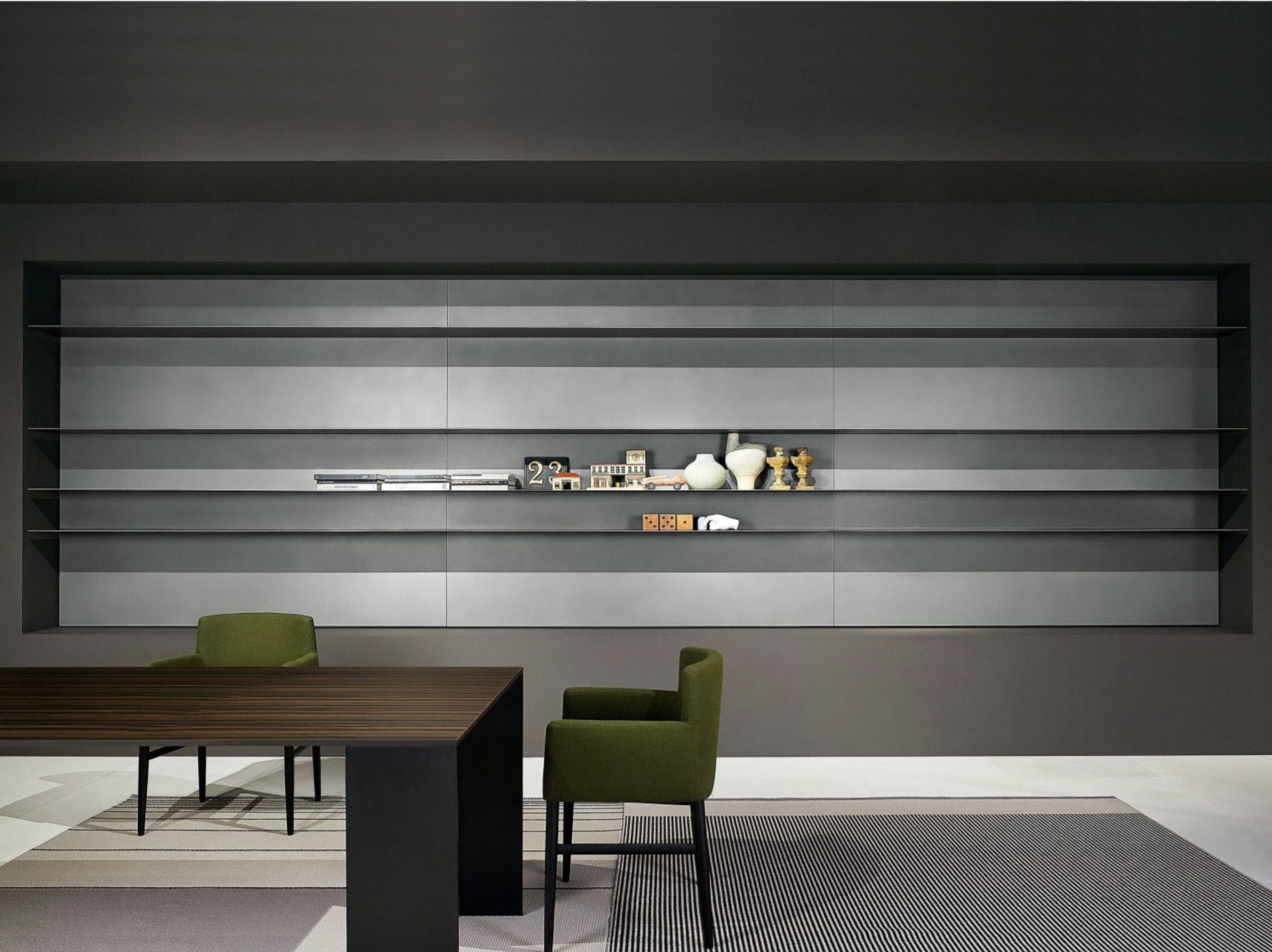 Modern minimalist room with a dark gray wall featuring built-in shelves, a dark wooden table, and two green chairs. The shelves hold small decorative objects including vases and trophies.
