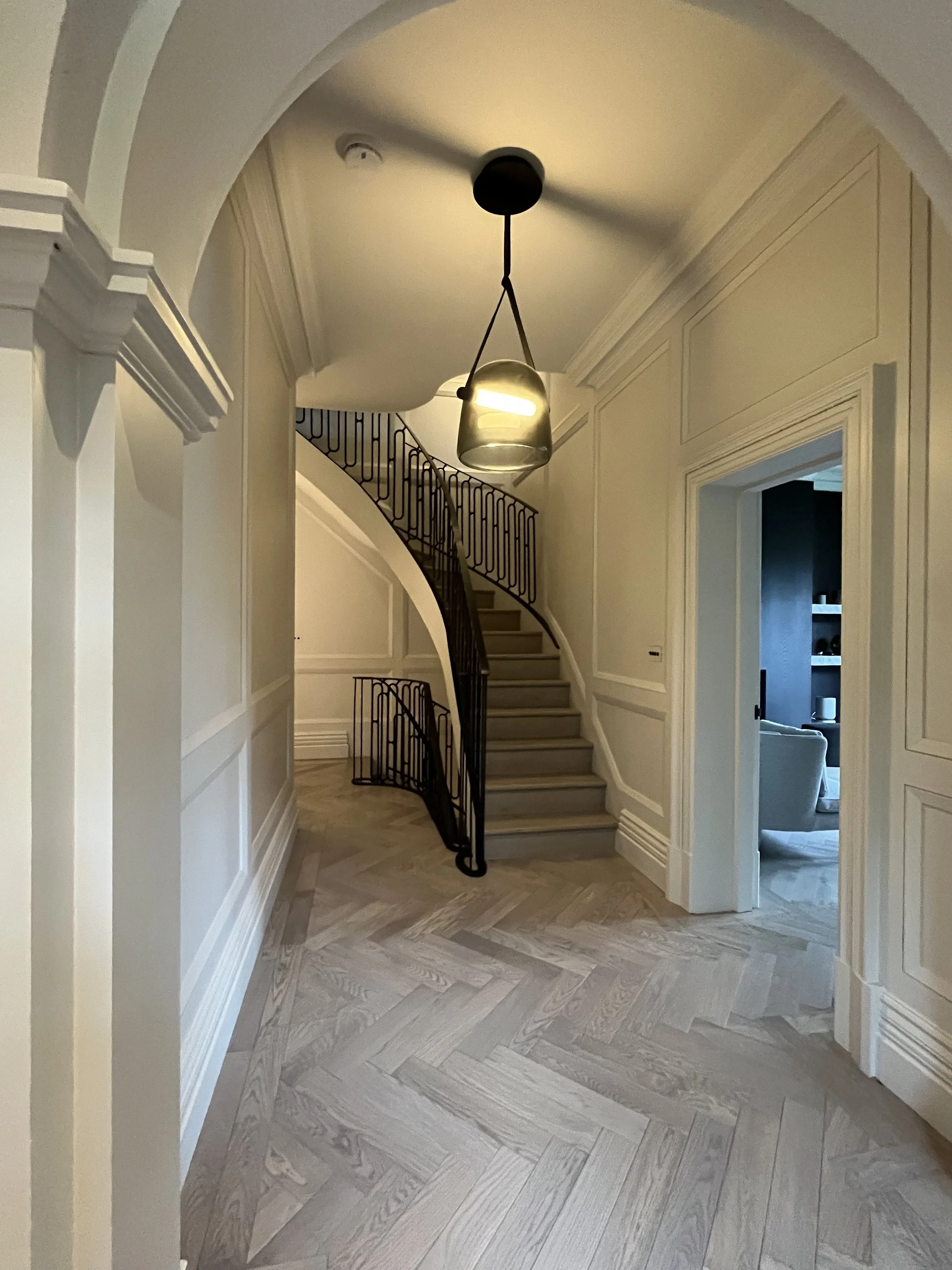 Interior view of a foyer with a curved staircase by Melides Design, modern hanging light, and light-colored wood parquet flooring. A doorway on the right leads to another room.