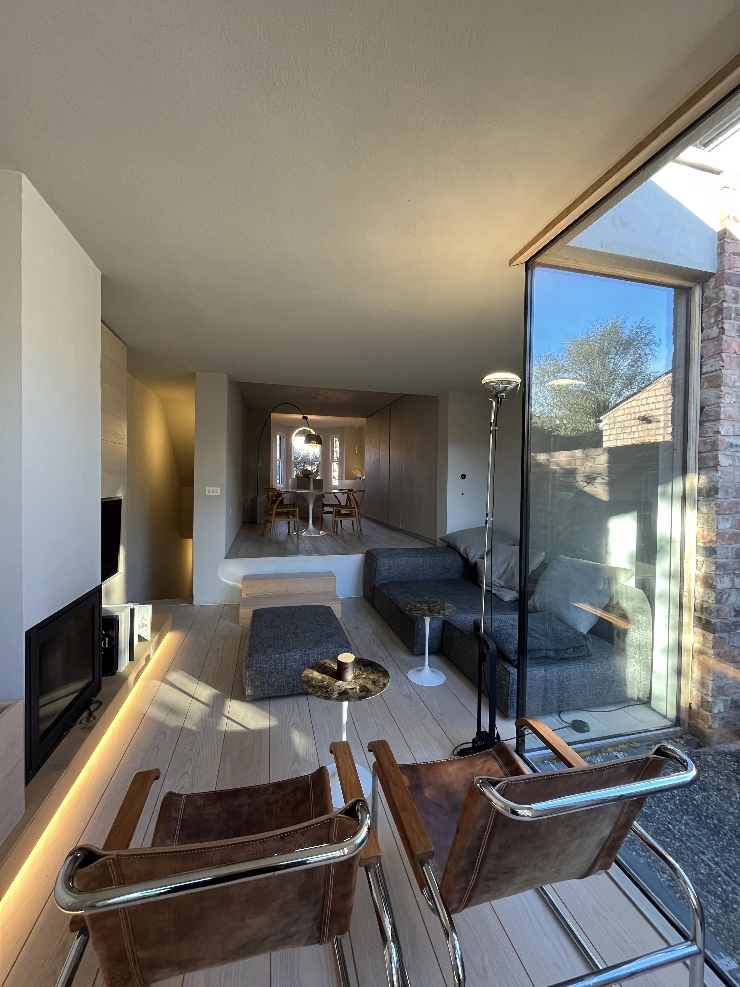Modern living room with a glass sliding door, a gray sectional sofa, two small marble-top side tables, a floor lamp, and a TV built into a white wall with light wood paneling.