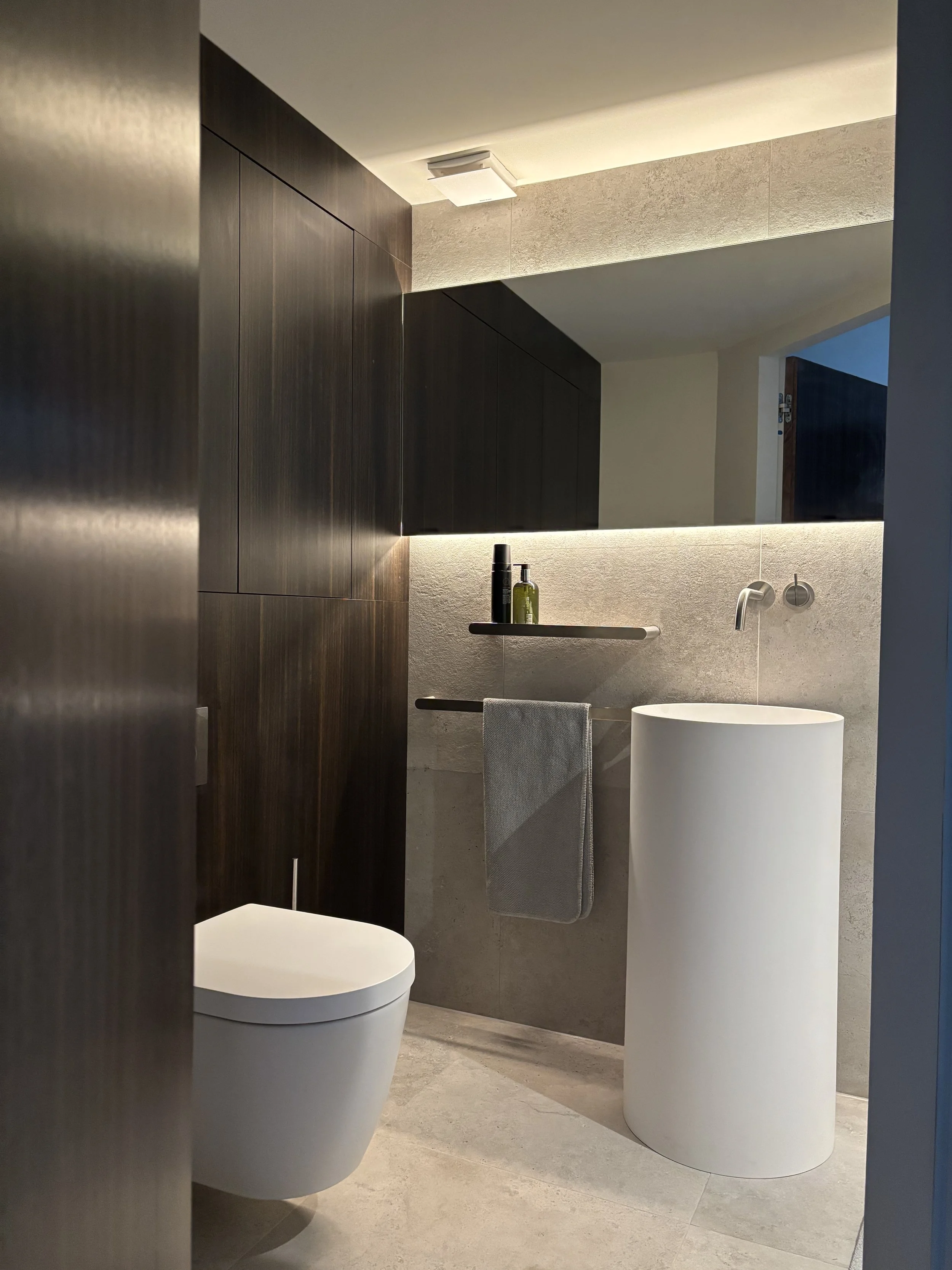 A modern bathroom featuring a white toilet, a freestanding white sink, dark wood cabinetry, a wall-mounted mirror, a floating shelf with toiletries, a towel hanging on a bar, modern lighting, and beige tiled walls and floor.