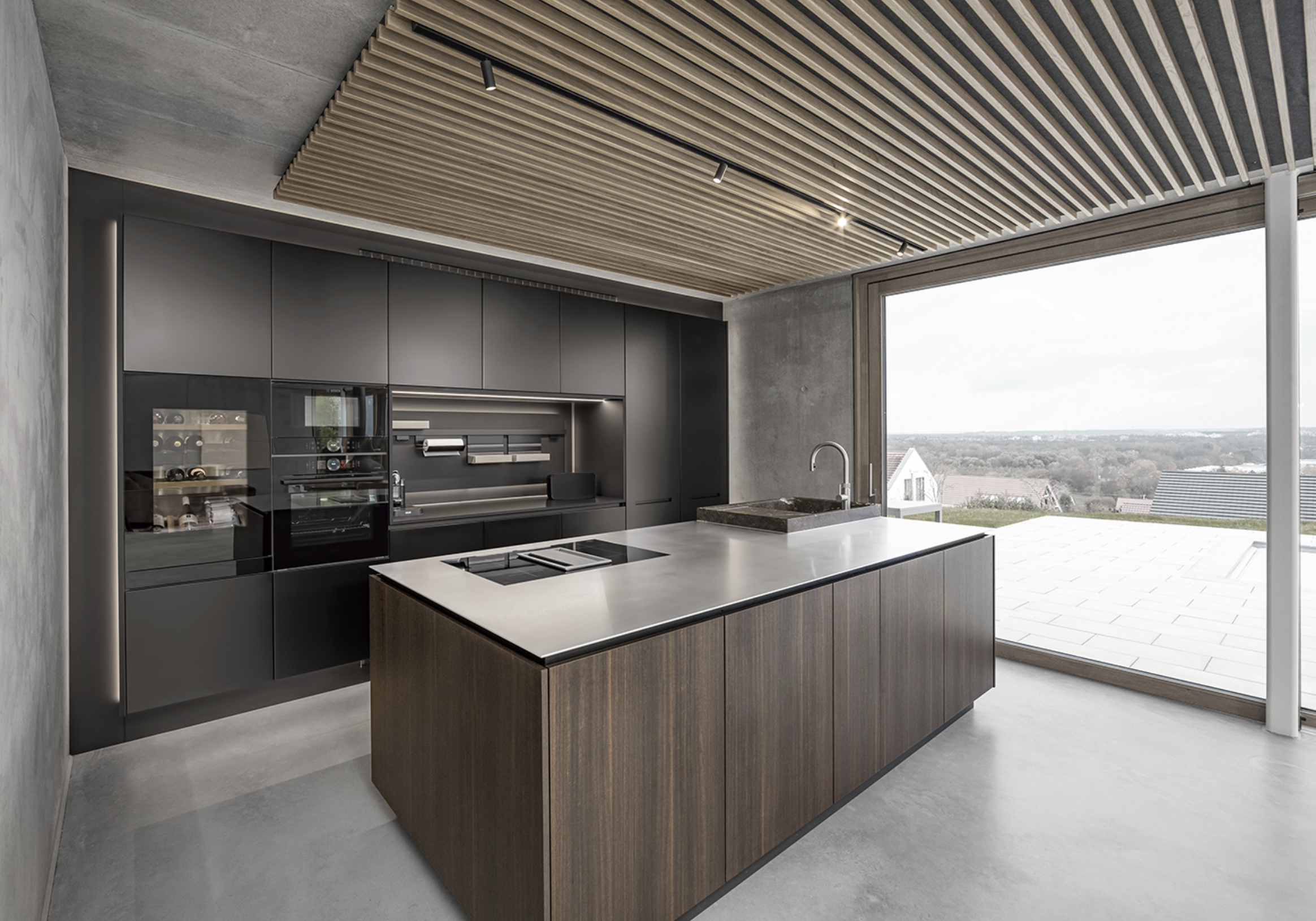 Modern kitchen with black cabinets, a large white island with a wooden base, a built-in oven, a stovetop, a sink with a window view, and wooden slatted ceiling.