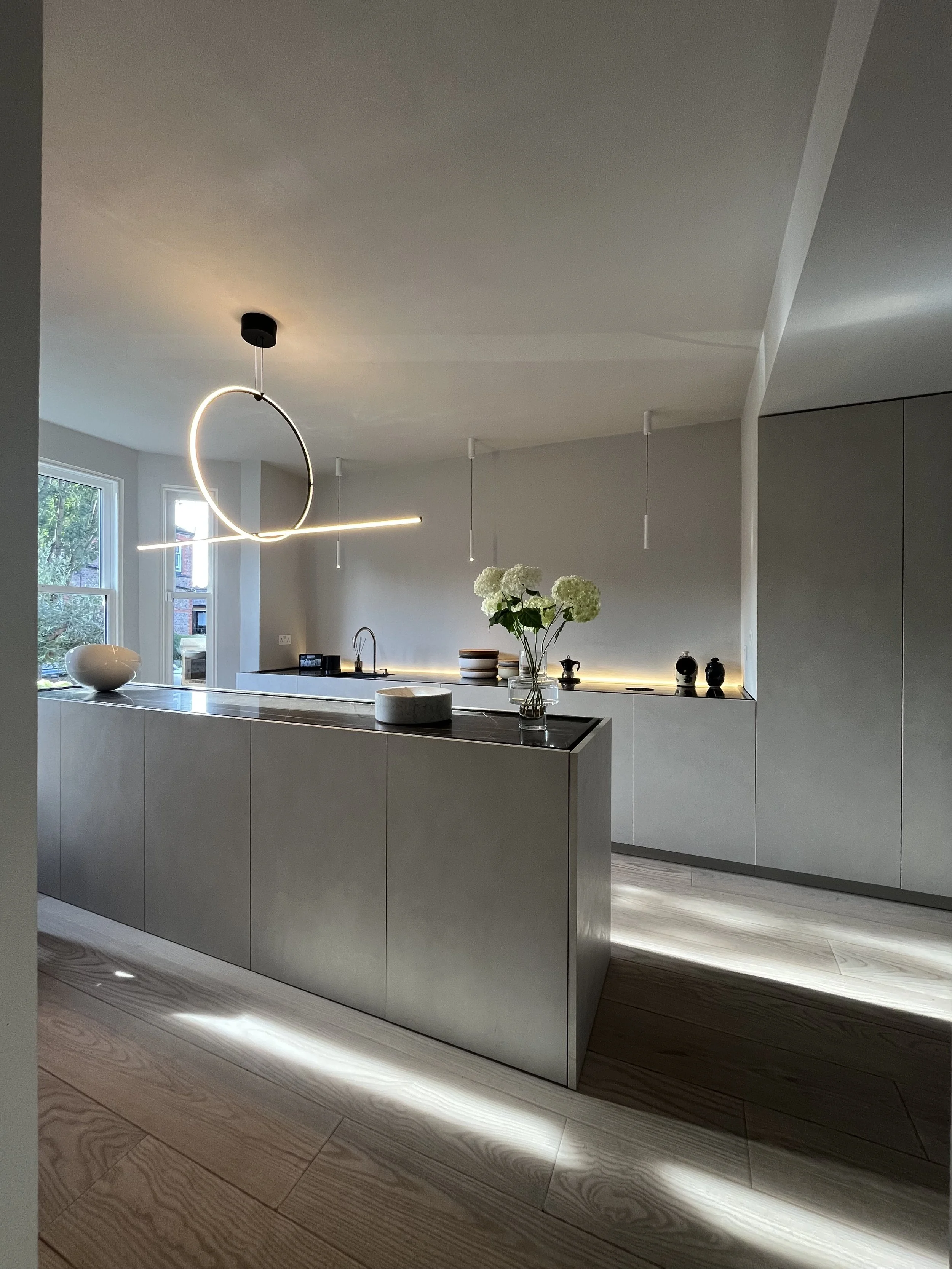 Modern minimalist kitchen with light-colored cabinetry, wooden flooring, vase of white flowers, and abstract hanging light fixture.
