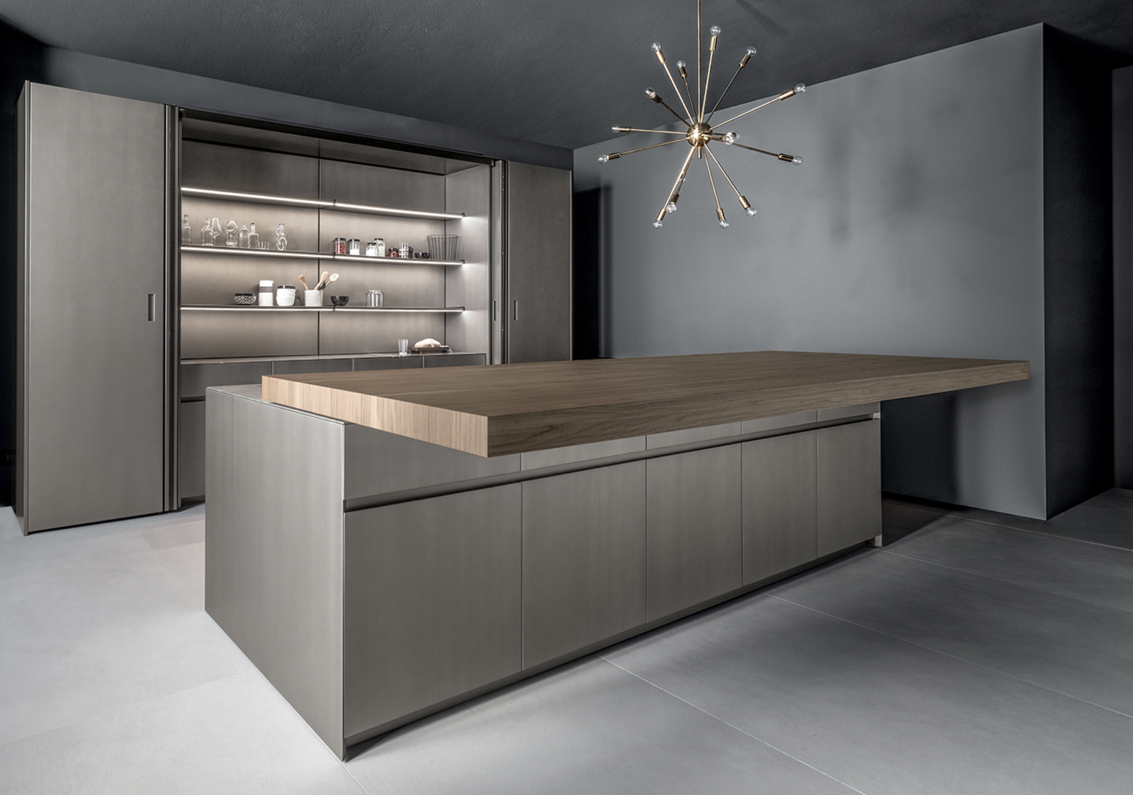Modern kitchen with minimalist gray cabinetry, a wooden countertop island, and a metallic shelving unit with dishes and glassware. A contemporary gold chandelier with exposed light bulbs hangs from the dark ceiling.