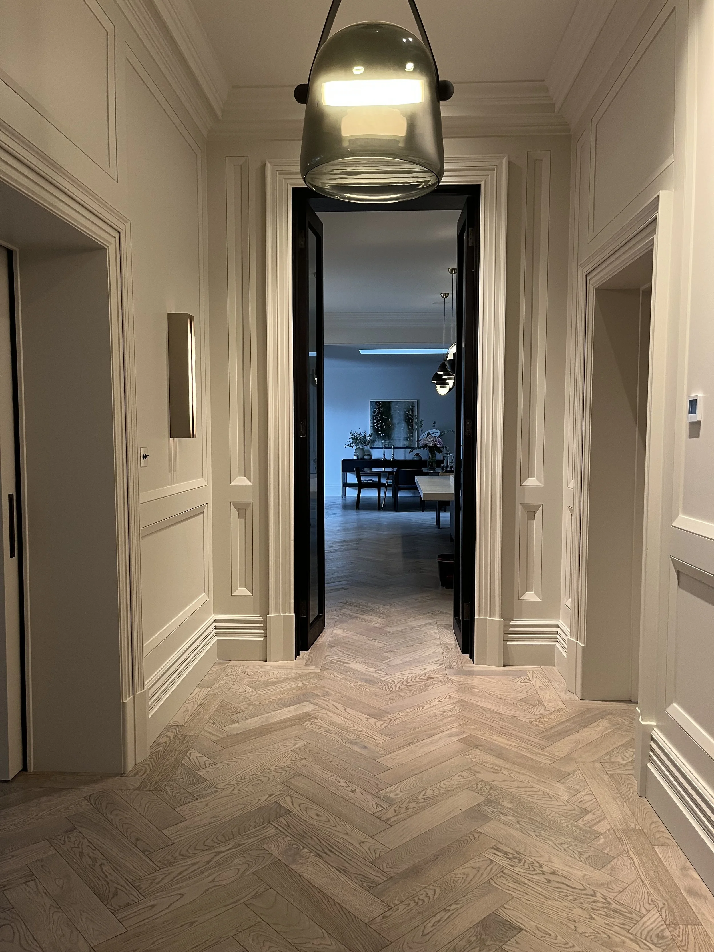 A hallway with beige walls, white crown molding, and a wooden herringbone floor leading to a dining room with a central table, chairs, and flowers.
