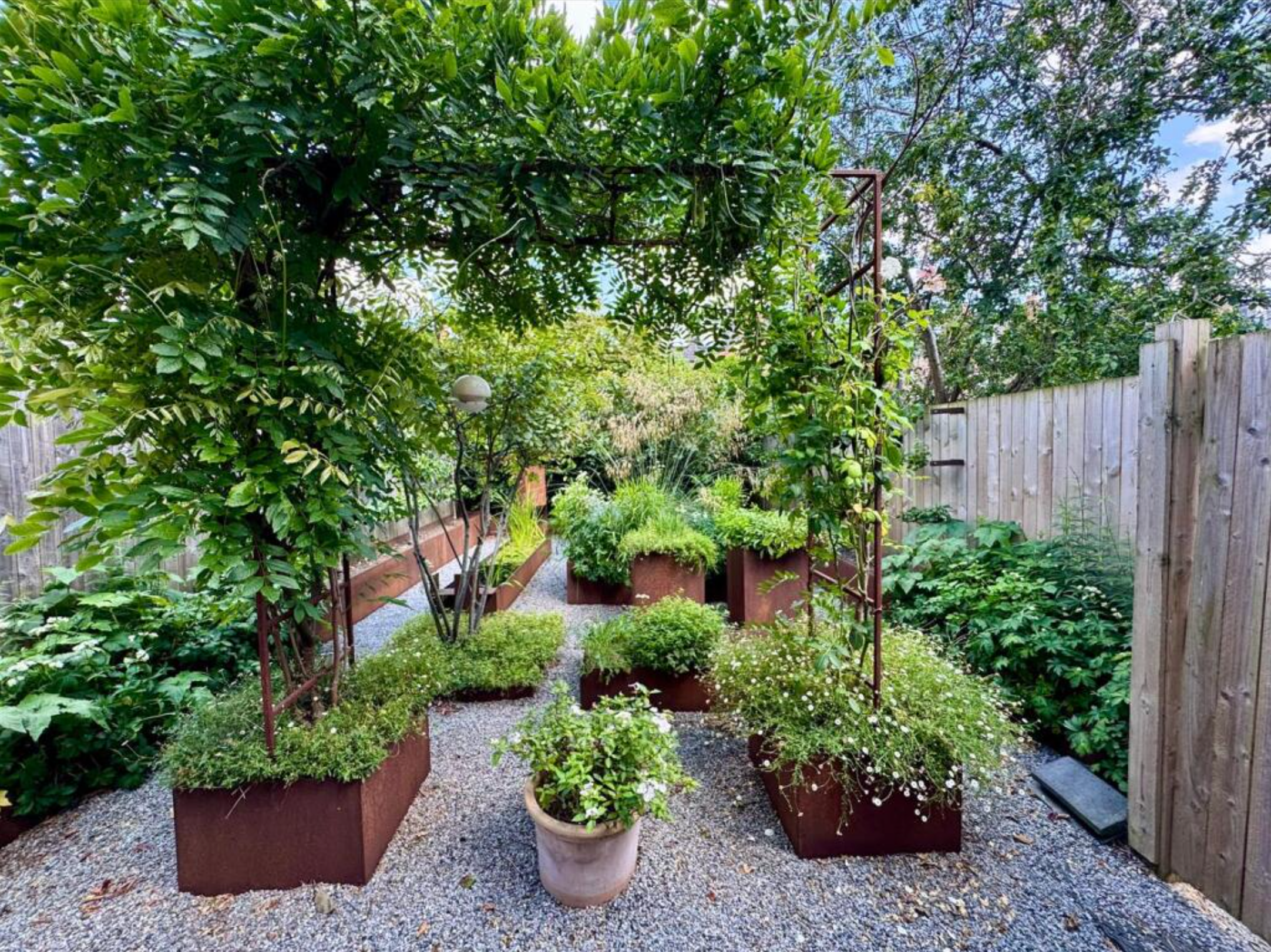 A small garden with potted plants and greenery, including trees, shrubs, and flower beds, enclosed by a wooden fence, with a gravel pathway and a metal archway.